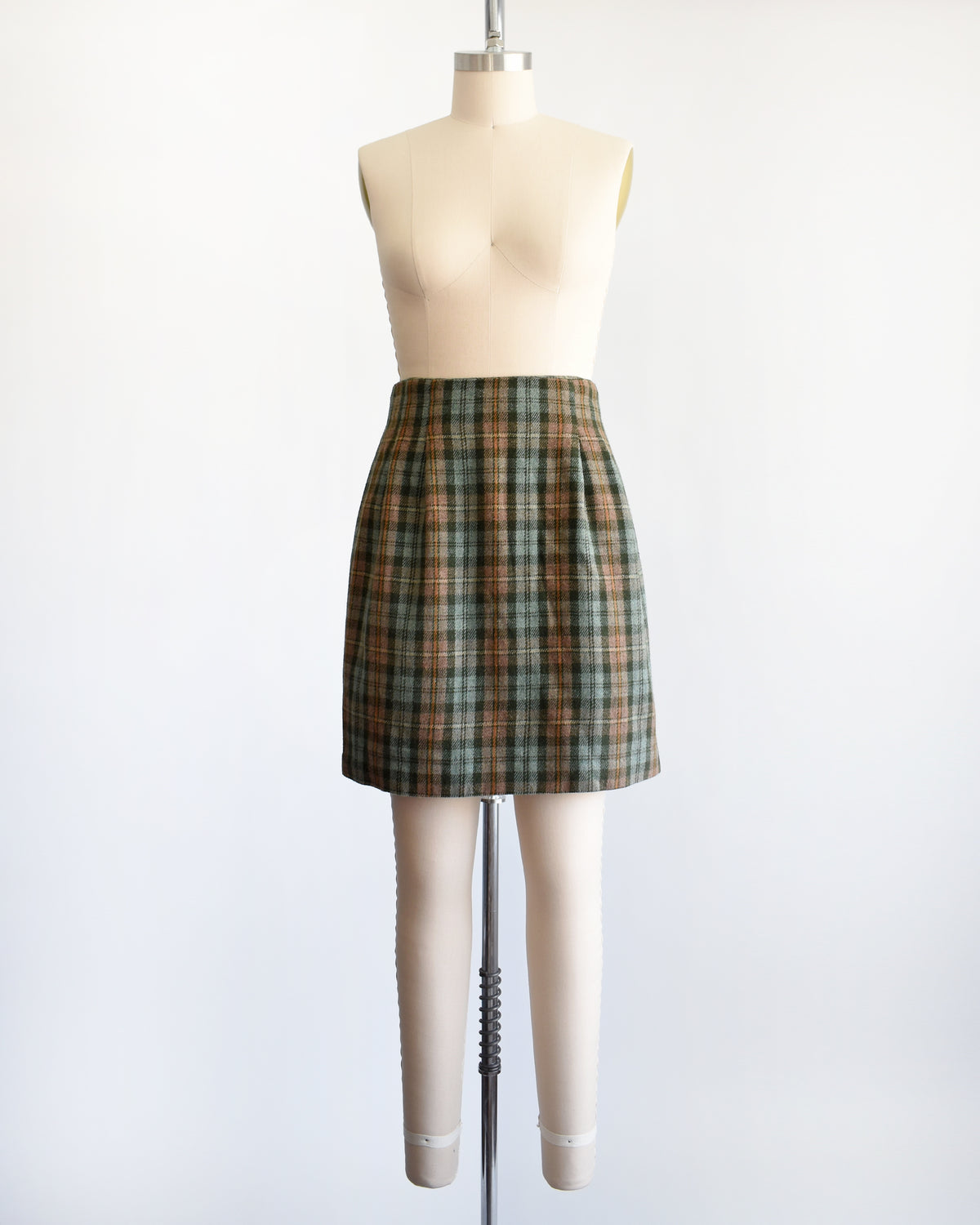 A vintage 1990s  green plaid mini skirt in green, brown, and beige tones, displayed on a cream-colored dress form mannequin against a plain white background.