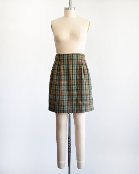 A vintage 1990s  green plaid mini skirt in green, brown, and beige tones, displayed on a cream-colored dress form mannequin against a plain white background.