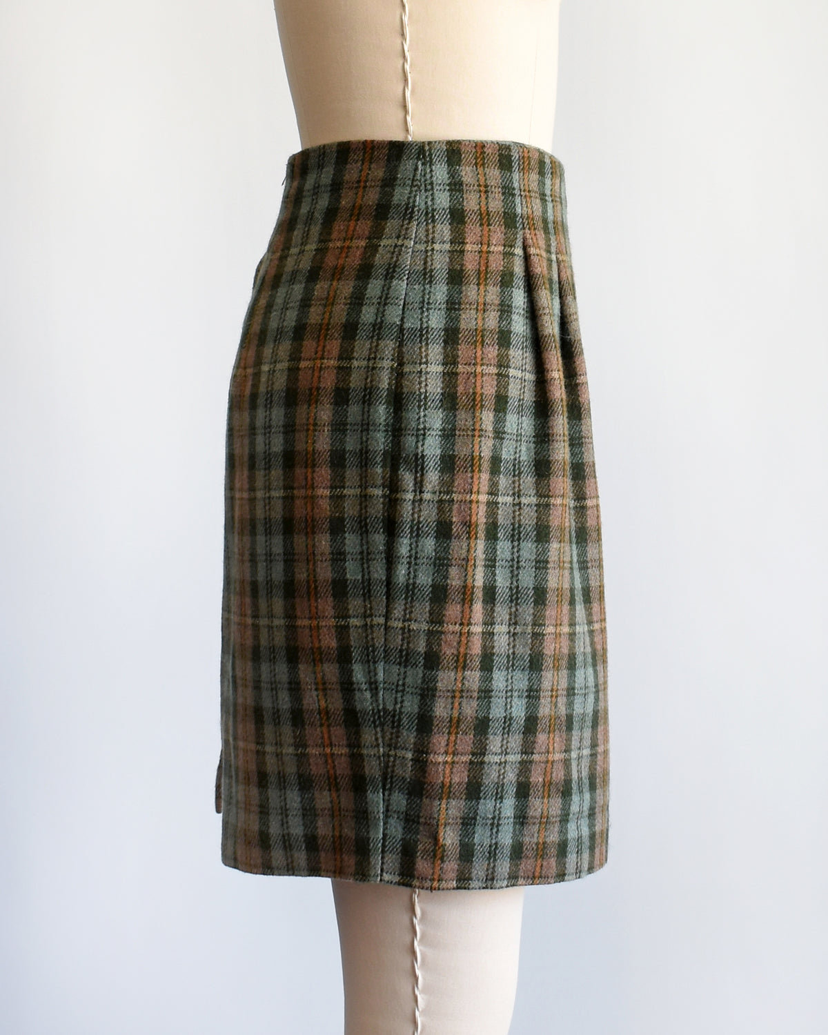 side view of a vintage 1990s  green plaid mini skirt in green, brown, and beige tones, displayed on a cream-colored dress form mannequin against a plain white background.