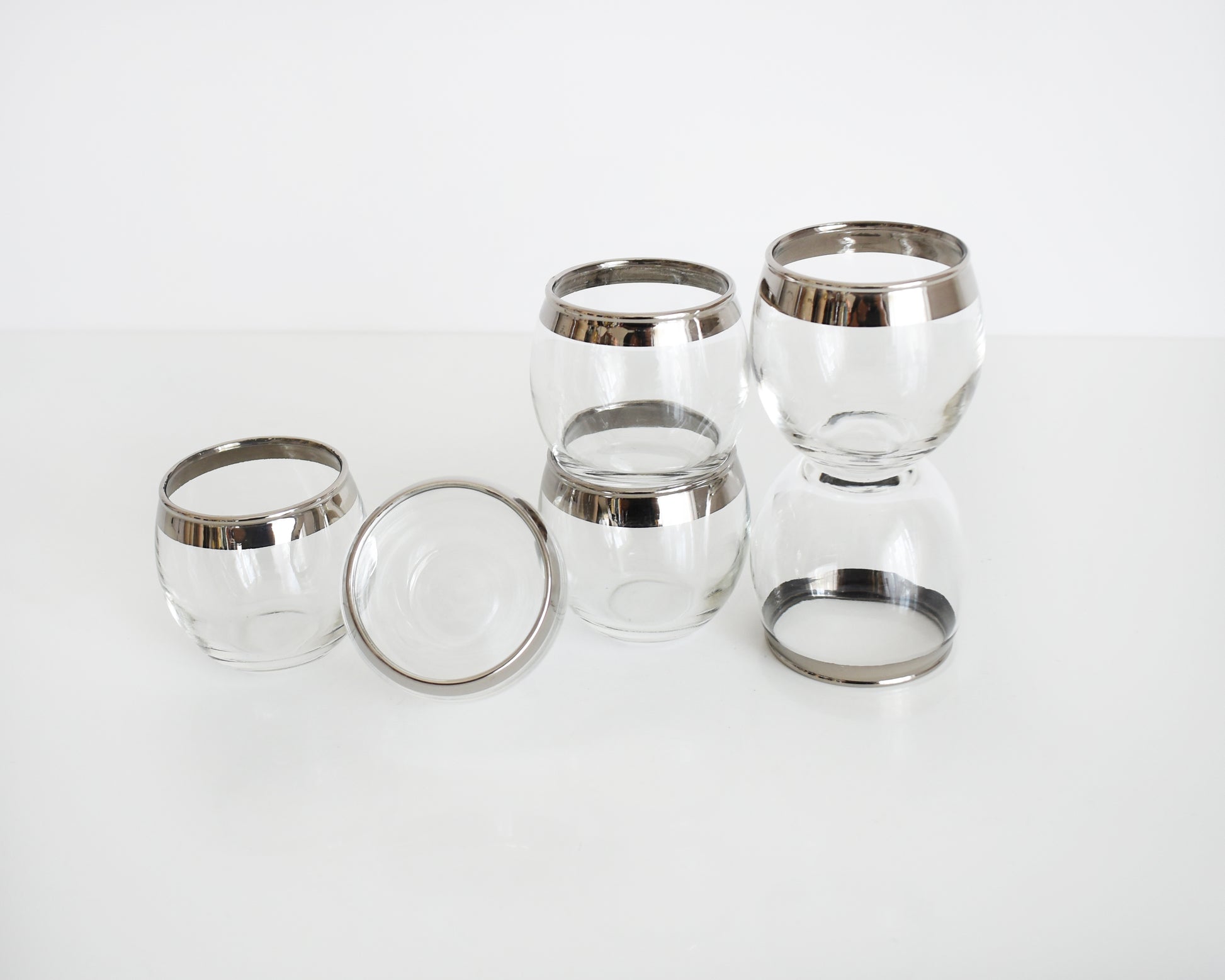 Six vintage silver rim roly poly shot glasses that have a ridge on the lip. One of the glasses is tipped over, one is stacked on top of another, and other is stacked with the glass underneath tipped over