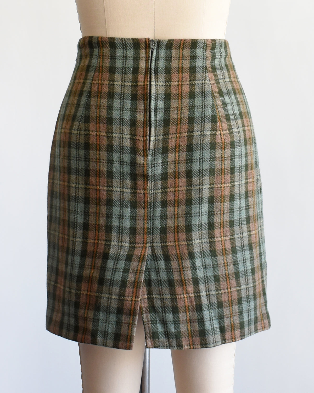 back view of a vintage 1990s  green plaid mini skirt in green, brown, and beige tones, displayed on a cream-colored dress form mannequin against a plain white background.