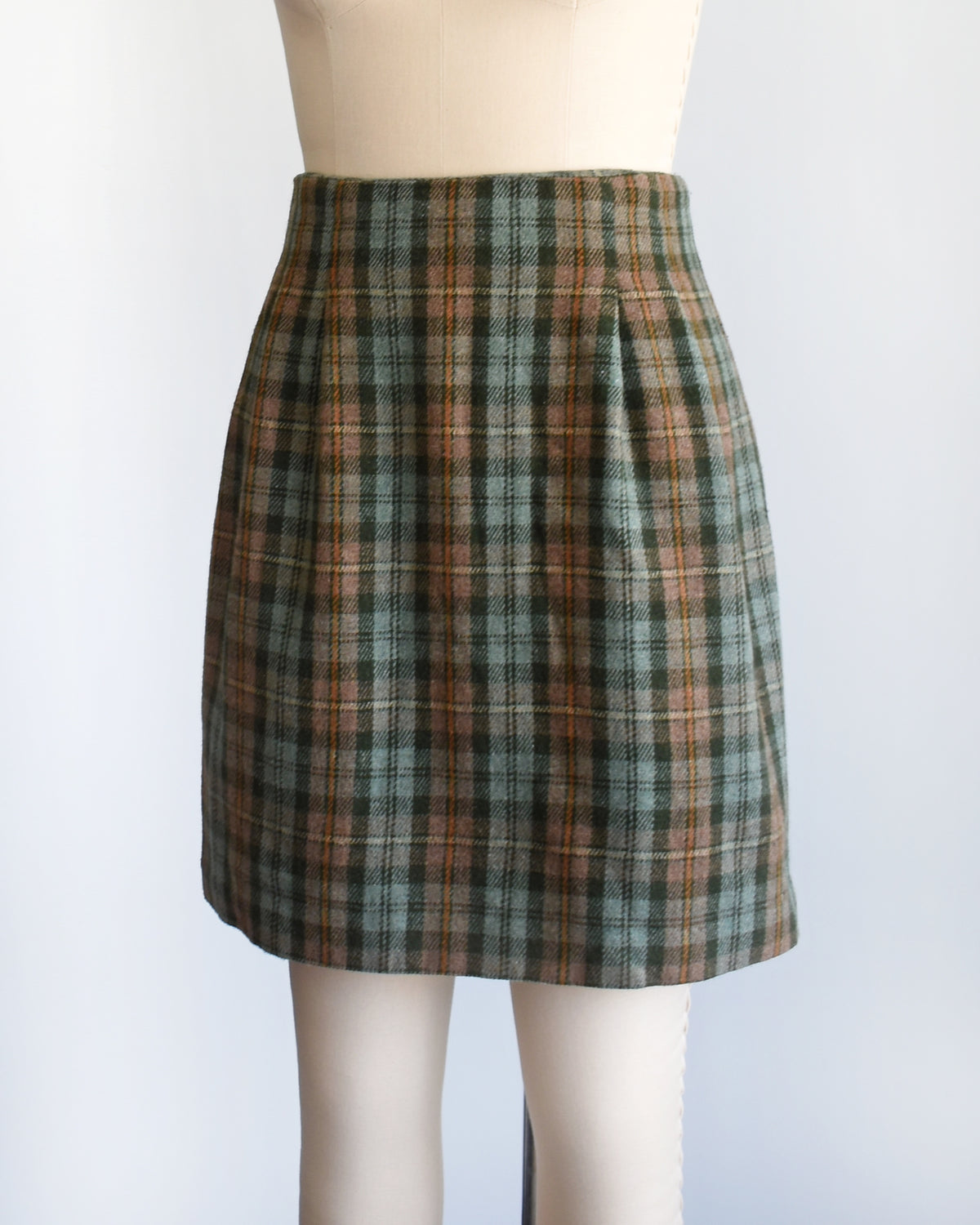 side front view of a vintage 1990s  green plaid mini skirt in green, brown, and beige tones, displayed on a cream-colored dress form mannequin against a plain white background.