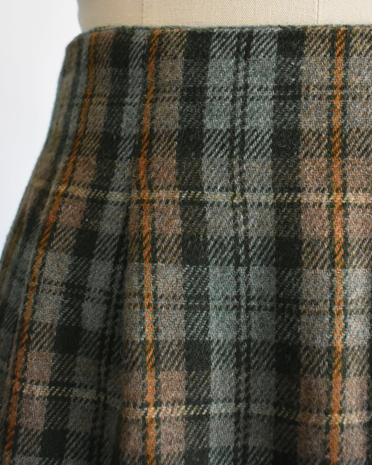 close up of a vintage 1990s  green plaid mini skirt in green, brown, and beige tones, displayed on a cream-colored dress form mannequin against a plain white background.