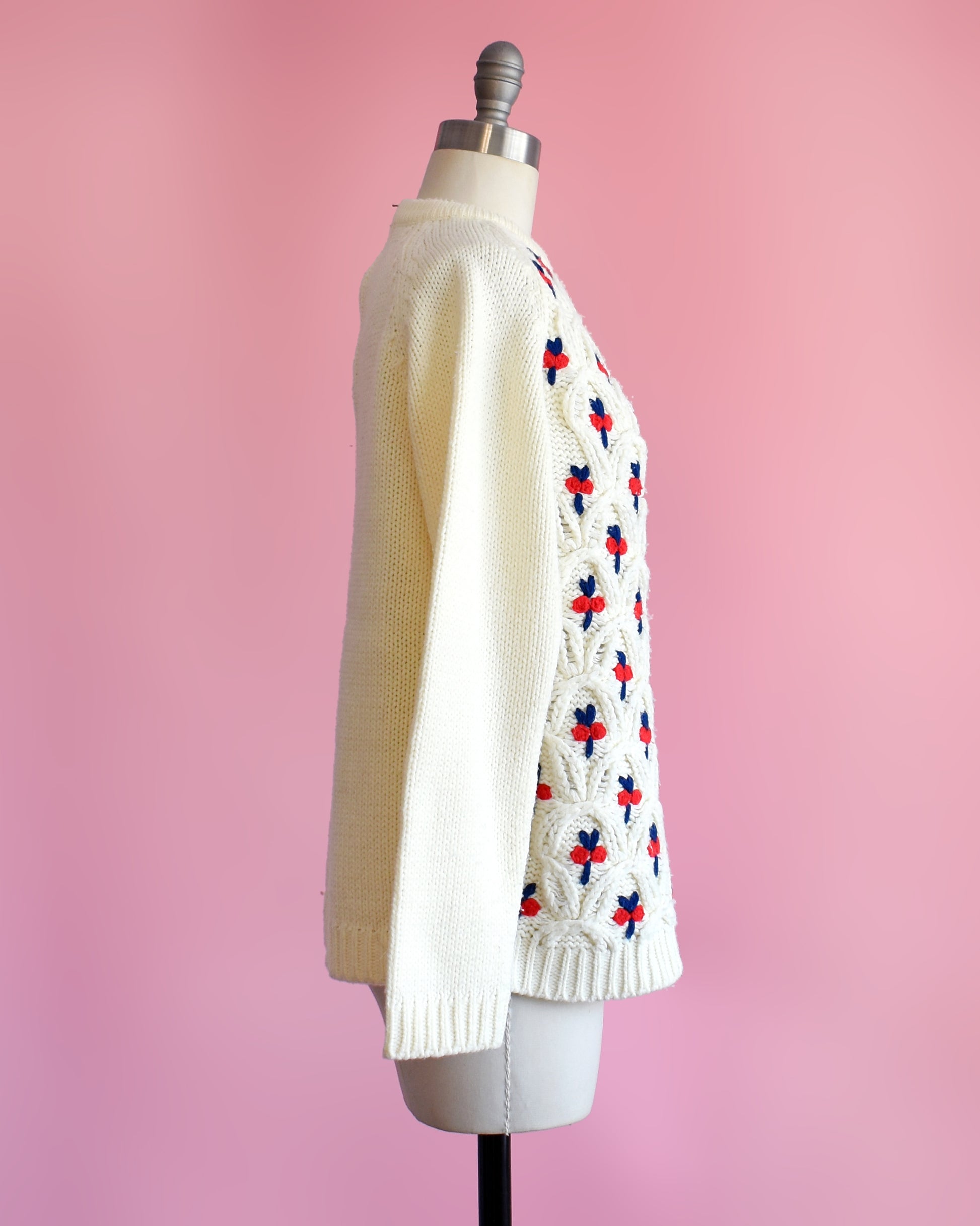 side view of a vintage 1970s white sweater with red and blue floral pattern on a mannequin against a pink background