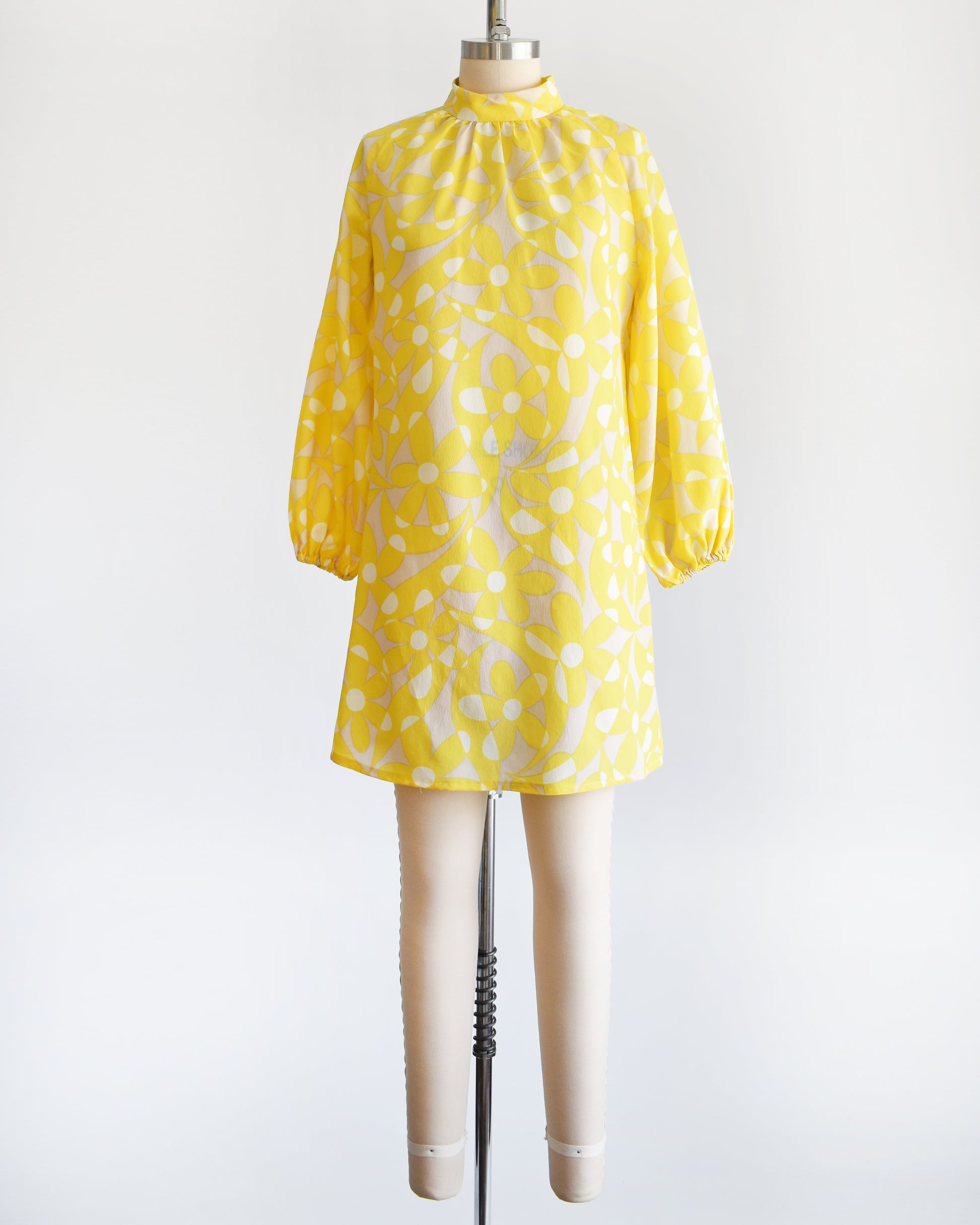 A vintage 1960s mod mini dress features a bright yellow and white floral pattern with swirls, set on a pale beige background. 
