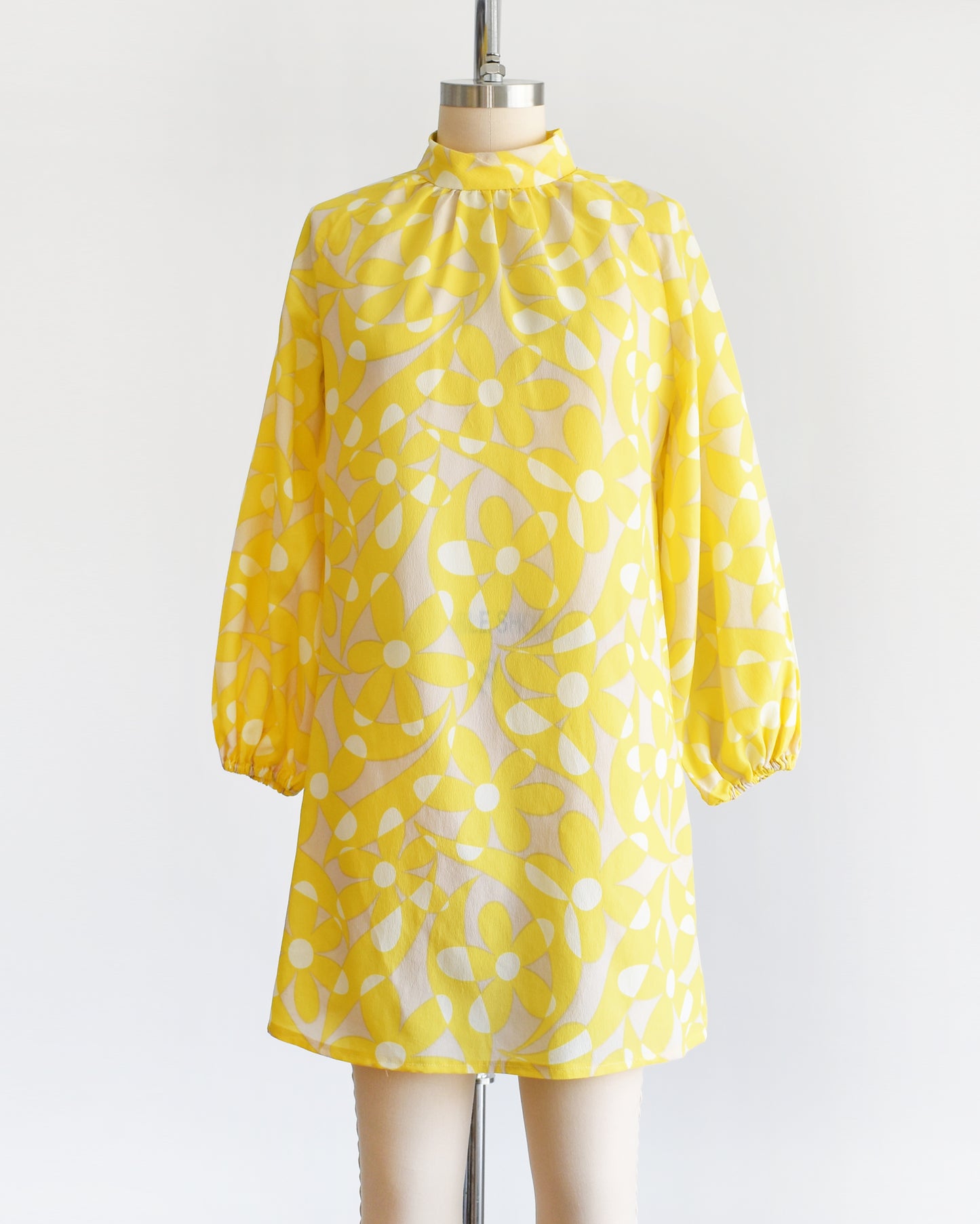 A vintage 1960s mod mini dress features a bright yellow and white floral pattern with swirls, set on a pale beige background. 