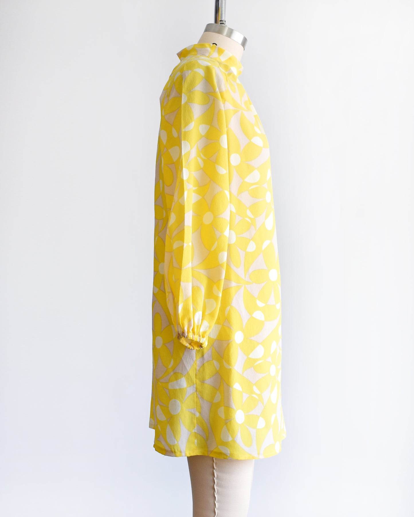 Side view of a vintage 1960s mod mini dress features a bright yellow and white floral pattern with swirls, set on a pale beige background. 