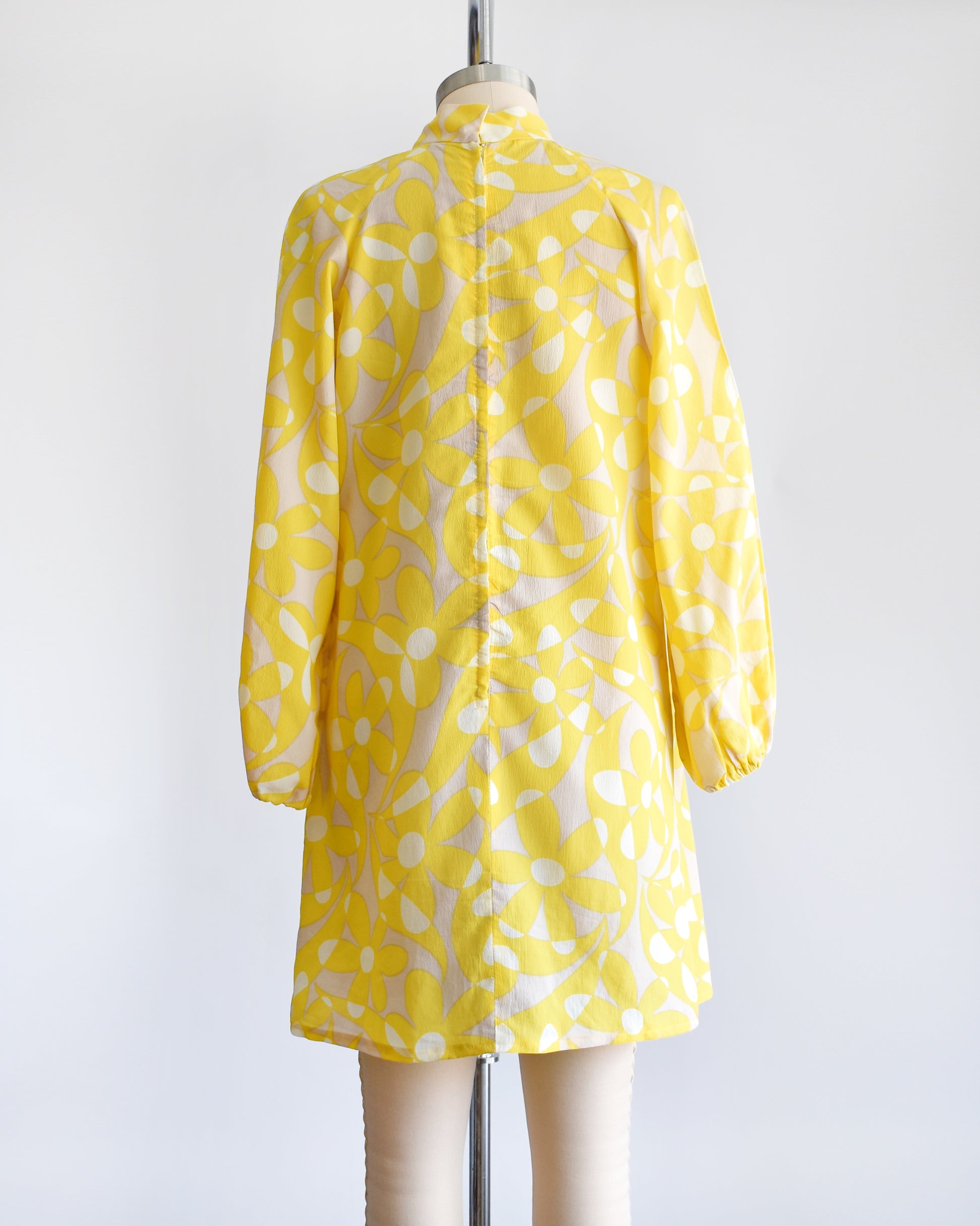 Back view if a vintage 1960s mod mini dress features a bright yellow and white floral pattern with swirls, set on a pale beige background. There's a zipper up the back