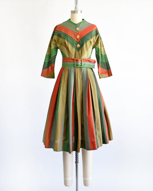 A vintage 1950s striped dress displayed on a mannequin against a white background. The dress features vertical stripes in green, orange, beige, and brown, with a matching waist belt and decorative buttons down the front bodice.