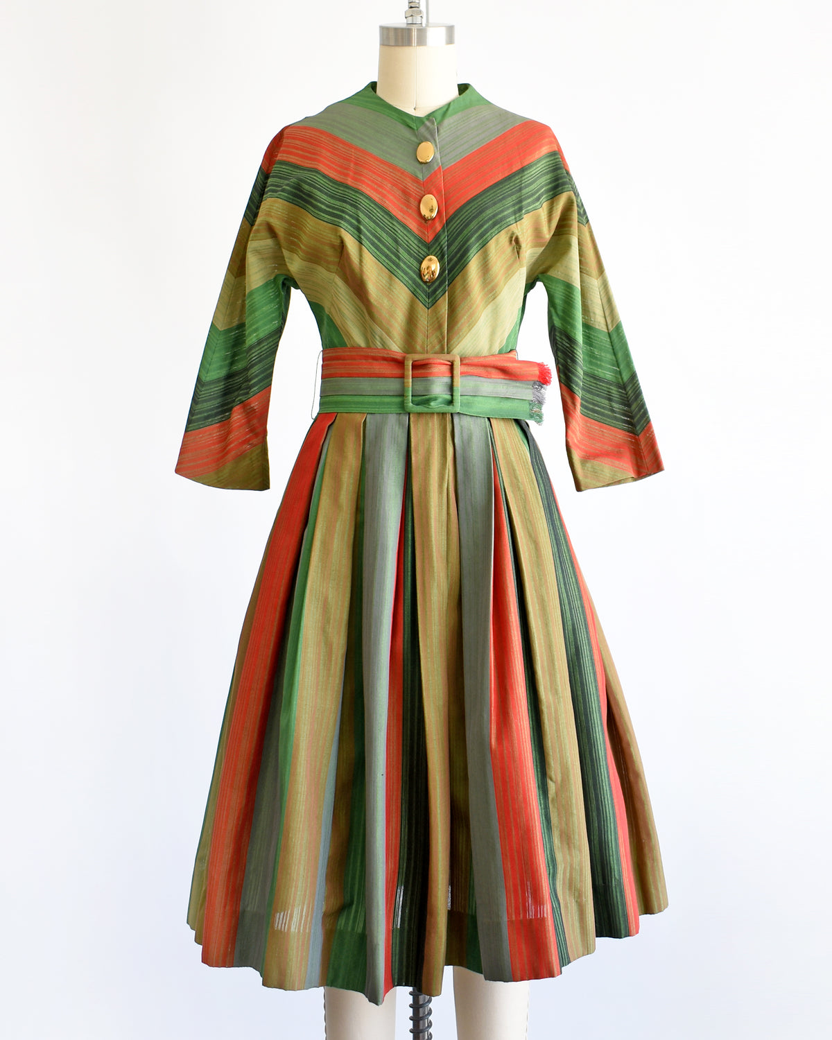 A vintage 1950s striped dress displayed on a mannequin against a white background. The dress features vertical stripes in green, orange, beige, and brown, with a matching waist belt and decorative buttons down the front bodice.