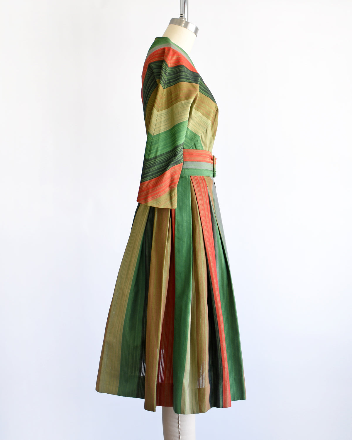side view of a vintage 1950s striped dress displayed on a mannequin against a white background. The dress features vertical stripes in green, orange, beige, and brown, with a matching waist belt and decorative buttons down the front bodice.