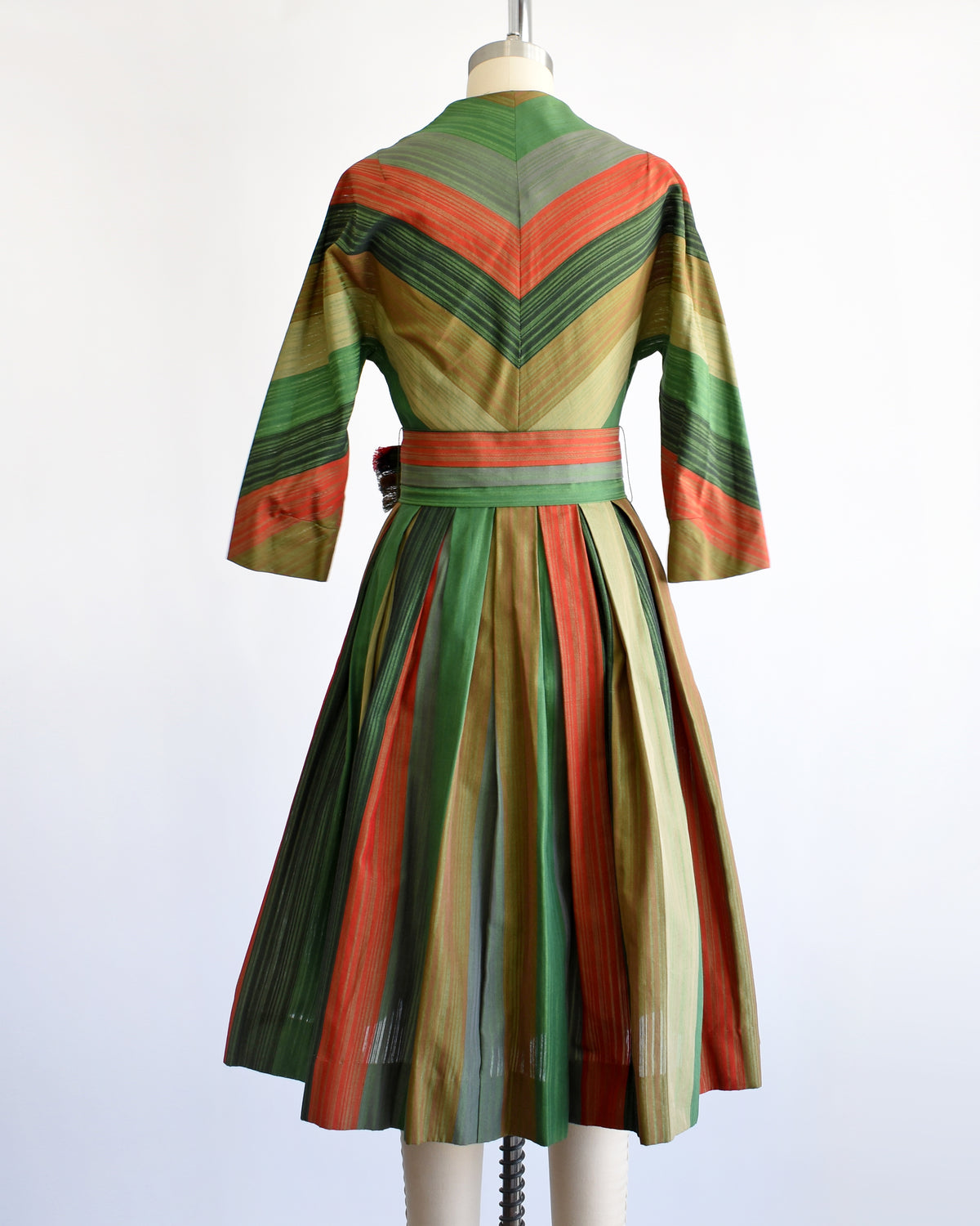 back view of a vintage 1950s striped dress displayed on a mannequin from the back, featuring a V-neck, three-quarter sleeves, a matching waist belt, and a pleated skirt in green, rust orange, beige, and brown stripes.