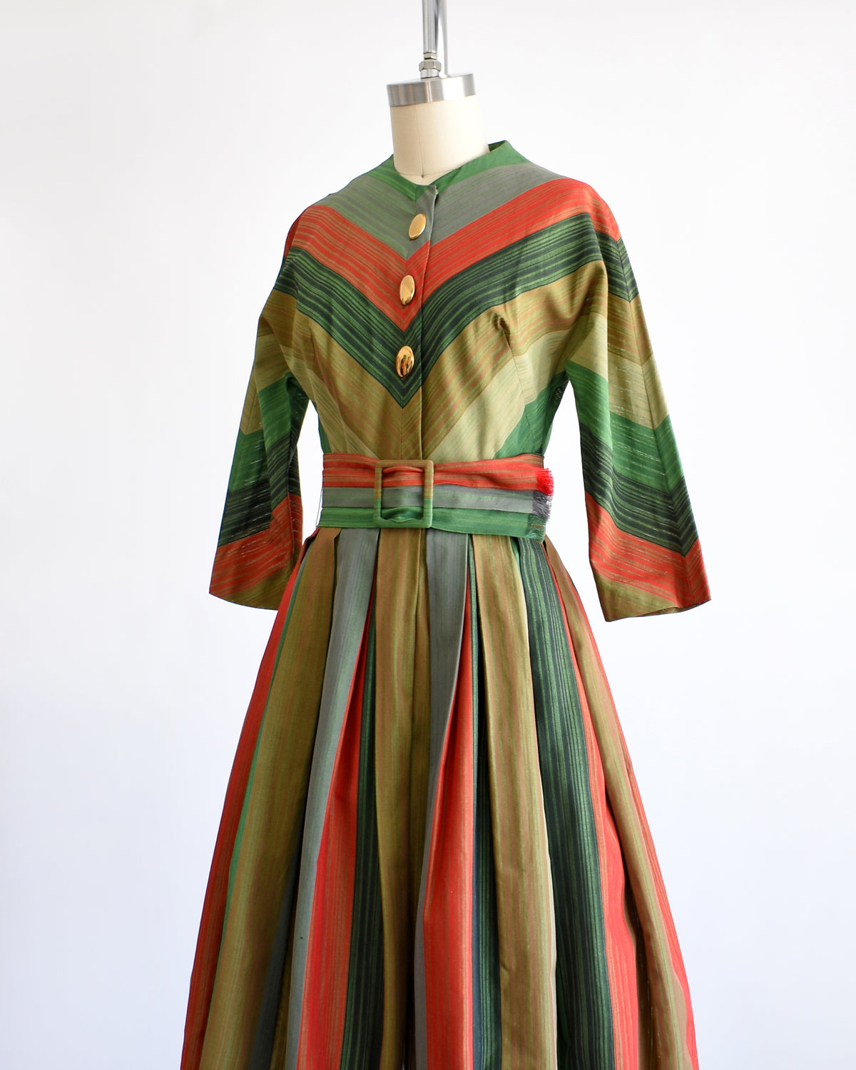 side view of a vintage 1950s striped dress displayed on a mannequin against a white background. The dress features vertical stripes in green, orange, beige, and brown, with a matching waist belt and decorative buttons down the front bodice.