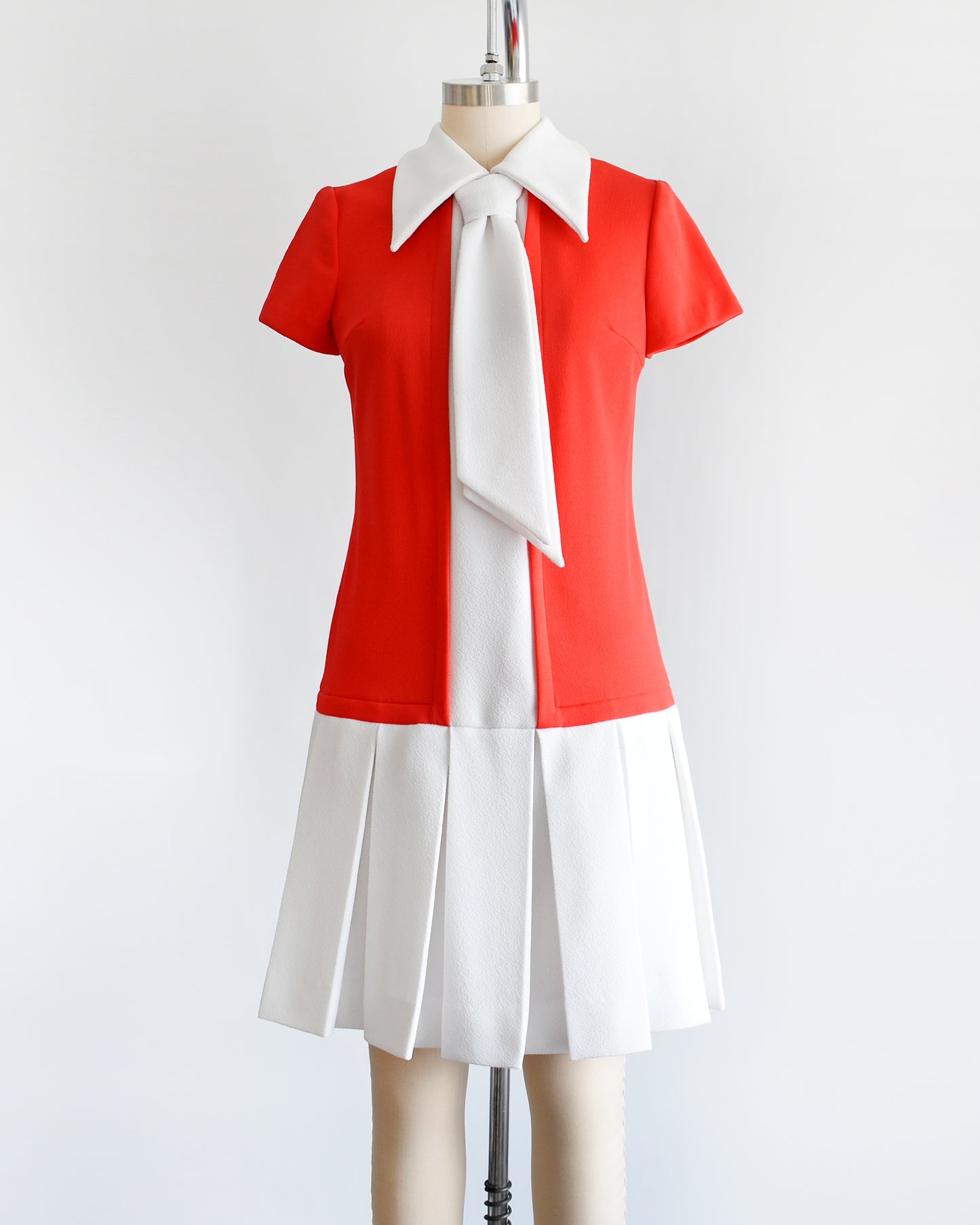 A vintage late 1960s early 1970s drop waist dress features a red bodice with a white collared neckline and ascot tie, accompanied by a white stripe down the front that flows into the white pleated skirt