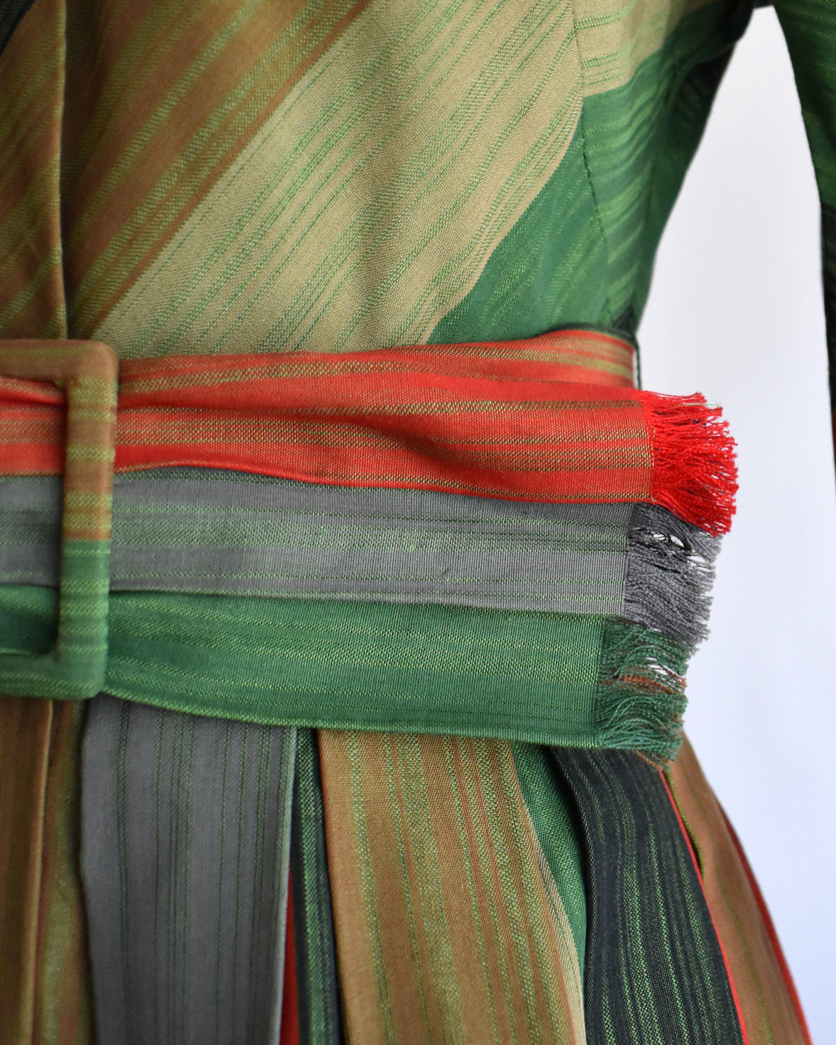 Close-up of the waist of the dress with a wide woven belt. The fabric features diagonal stripes in green, tan, and brown, with a prominent red-orange stripe across the belt and a fringed end.