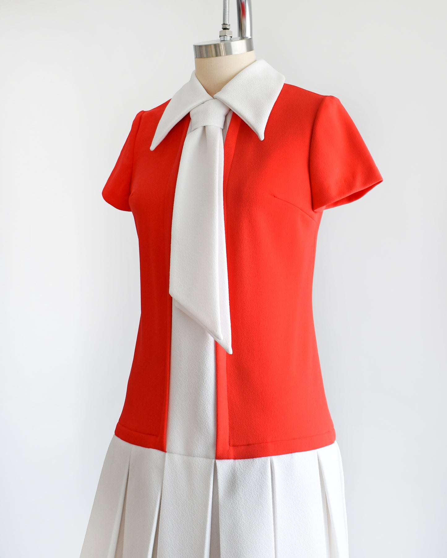 Side front view of a vintage late 1960s early 1970s drop waist dress features a red bodice with a white collared neckline and ascot tie, accompanied by a white stripe down the front that flows into the white pleated skirt