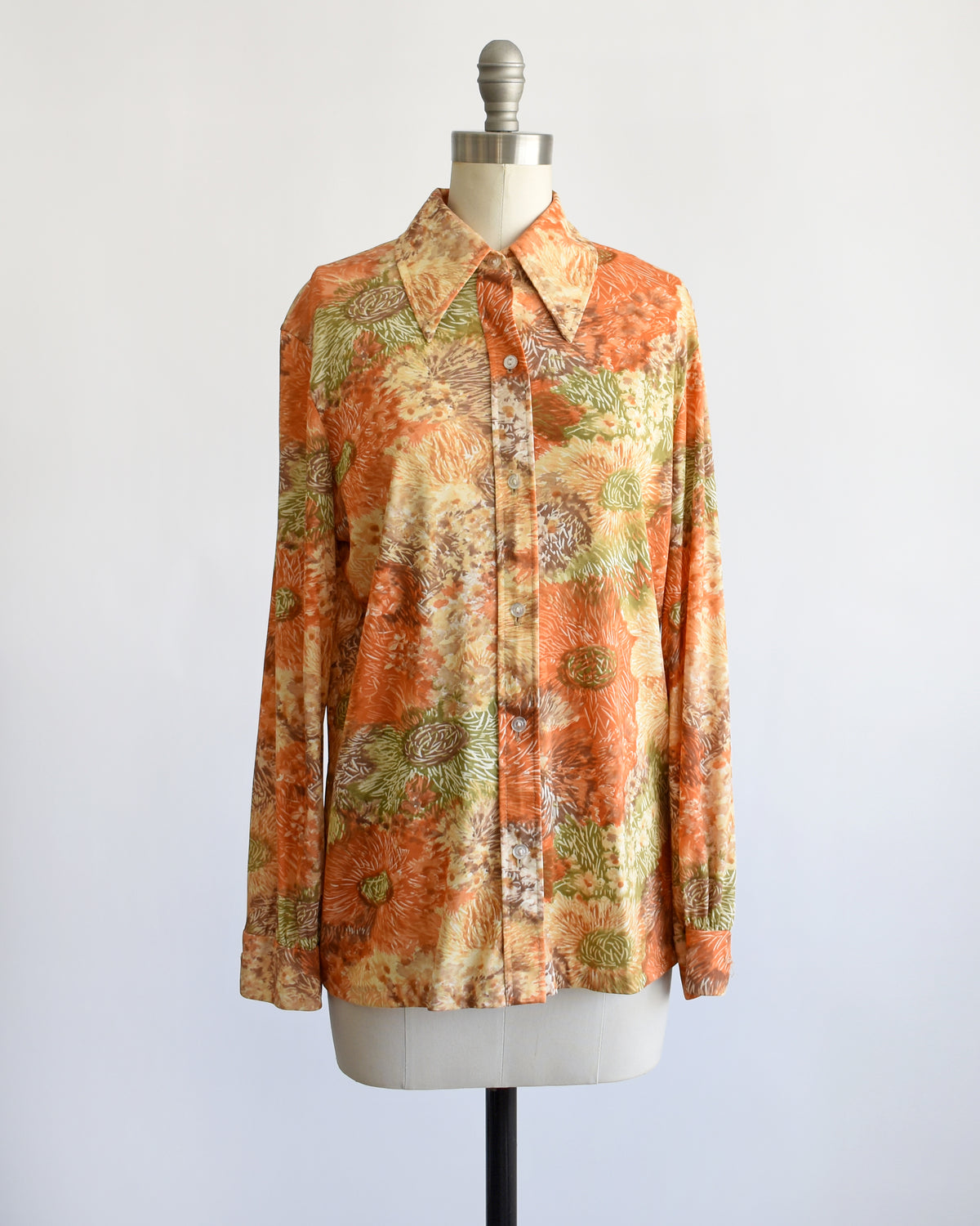 a vintage 1970s long-sleeve button-down blouse which features an abstract floral watercolor print in orange, rust, and olive green tones displayed on a dress form.
