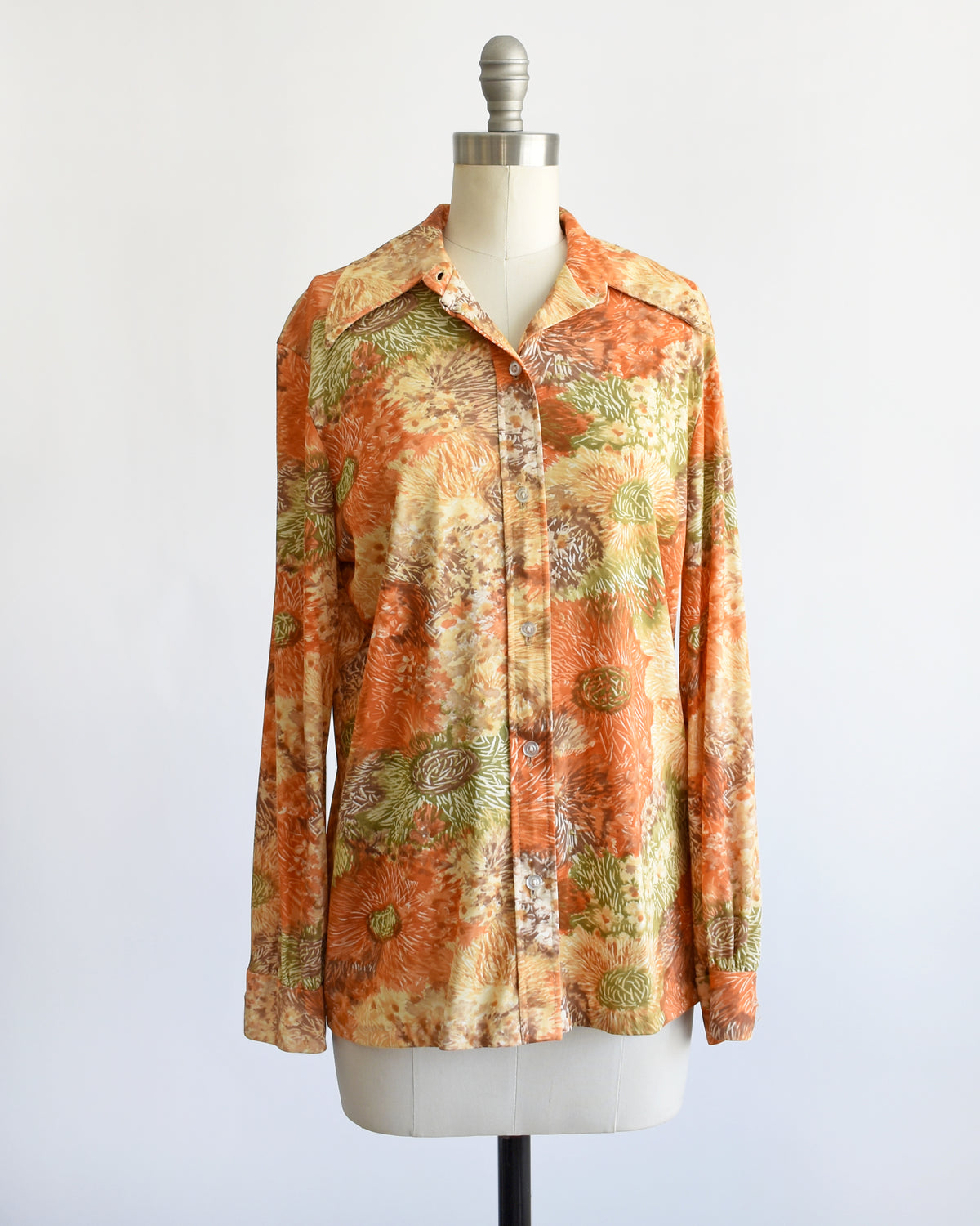 a vintage 1970s long-sleeve button-down blouse which features an abstract floral watercolor print in orange, rust, and olive green tones displayed on a dress form. the top button is unbuttoned.