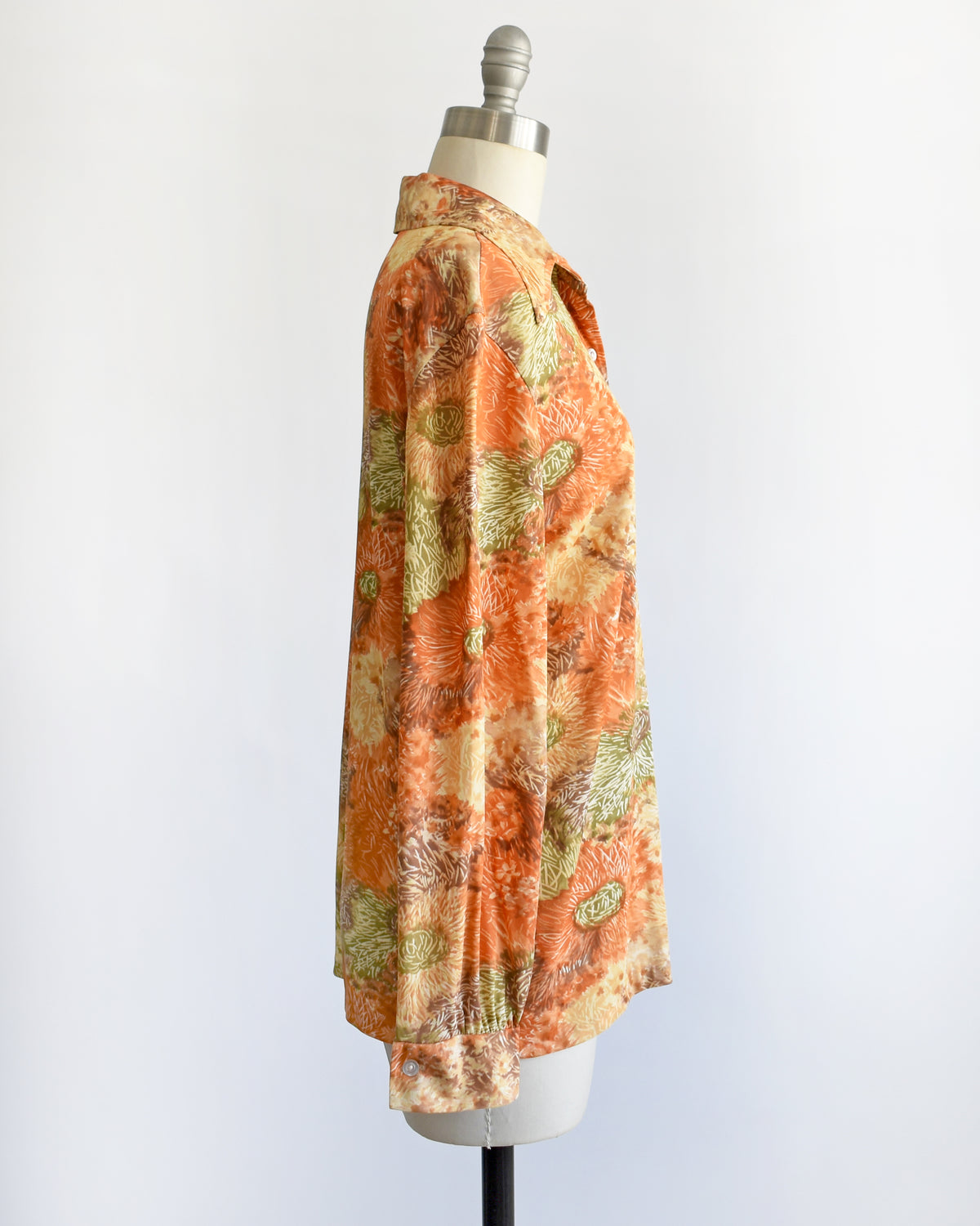 a vintage 1970s long-sleeve button-down blouse which features an abstract floral watercolor print in orange, rust, and olive green tones displayed on a dress form.