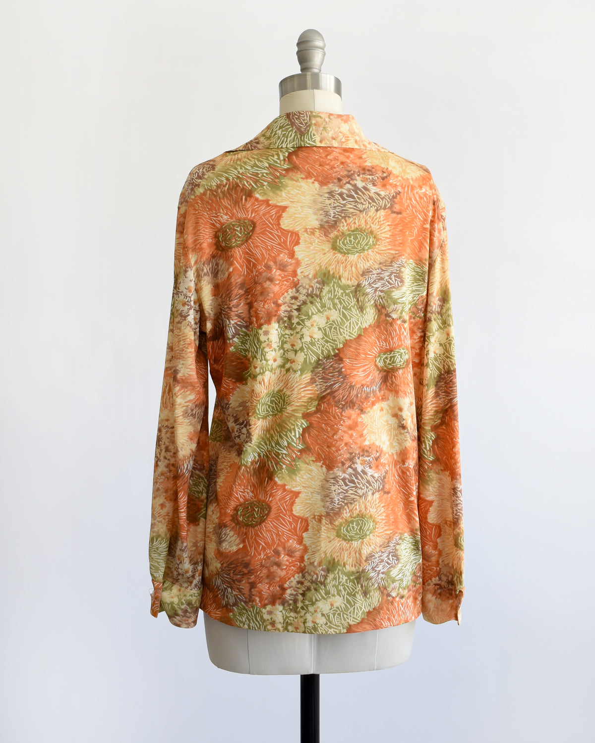 a vintage 1970s long-sleeve button-down blouse which features an abstract floral watercolor print in orange, rust, and olive green tones displayed on a dress form.
