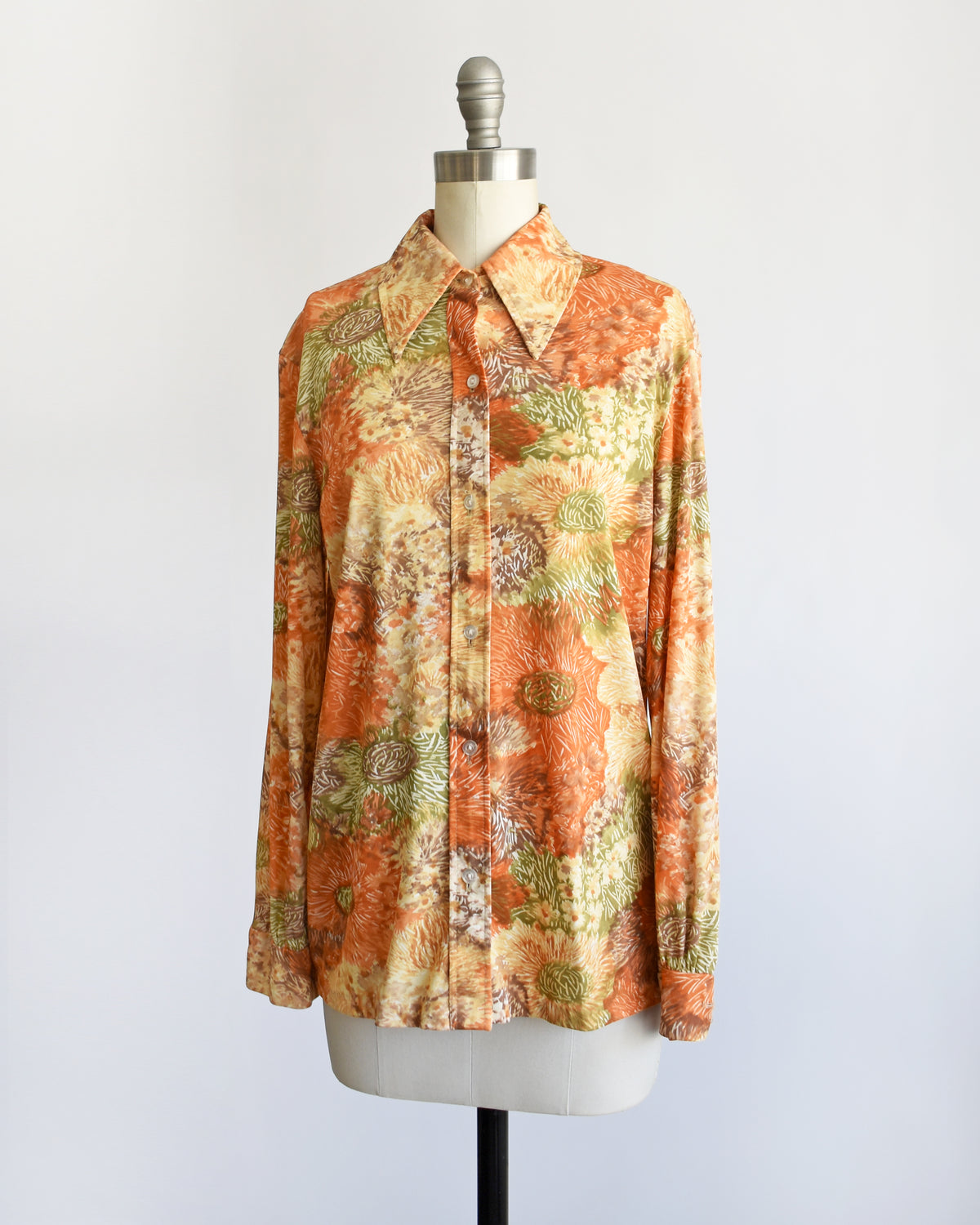 a vintage 1970s long-sleeve button-down blouse which features an abstract floral watercolor print in orange, rust, and olive green tones displayed on a dress form.