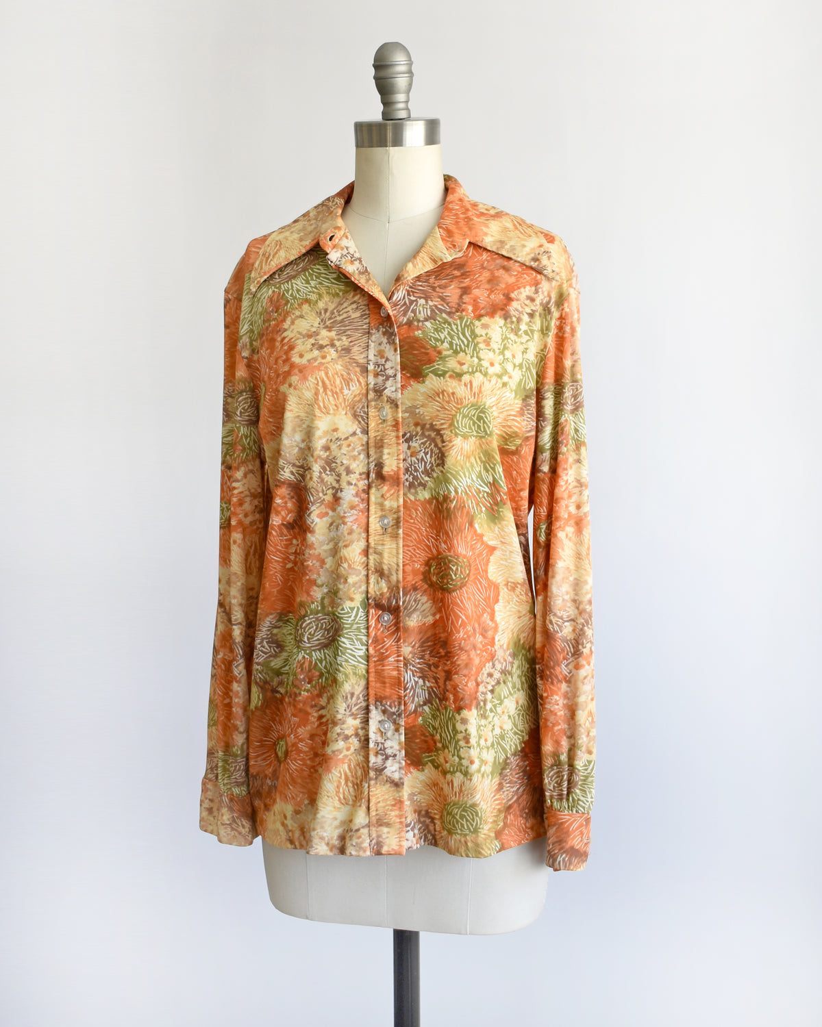 a vintage 1970s long-sleeve button-down blouse which features an abstract floral watercolor print in orange, rust, and olive green tones displayed on a dress form. the top button is unbuttoned.