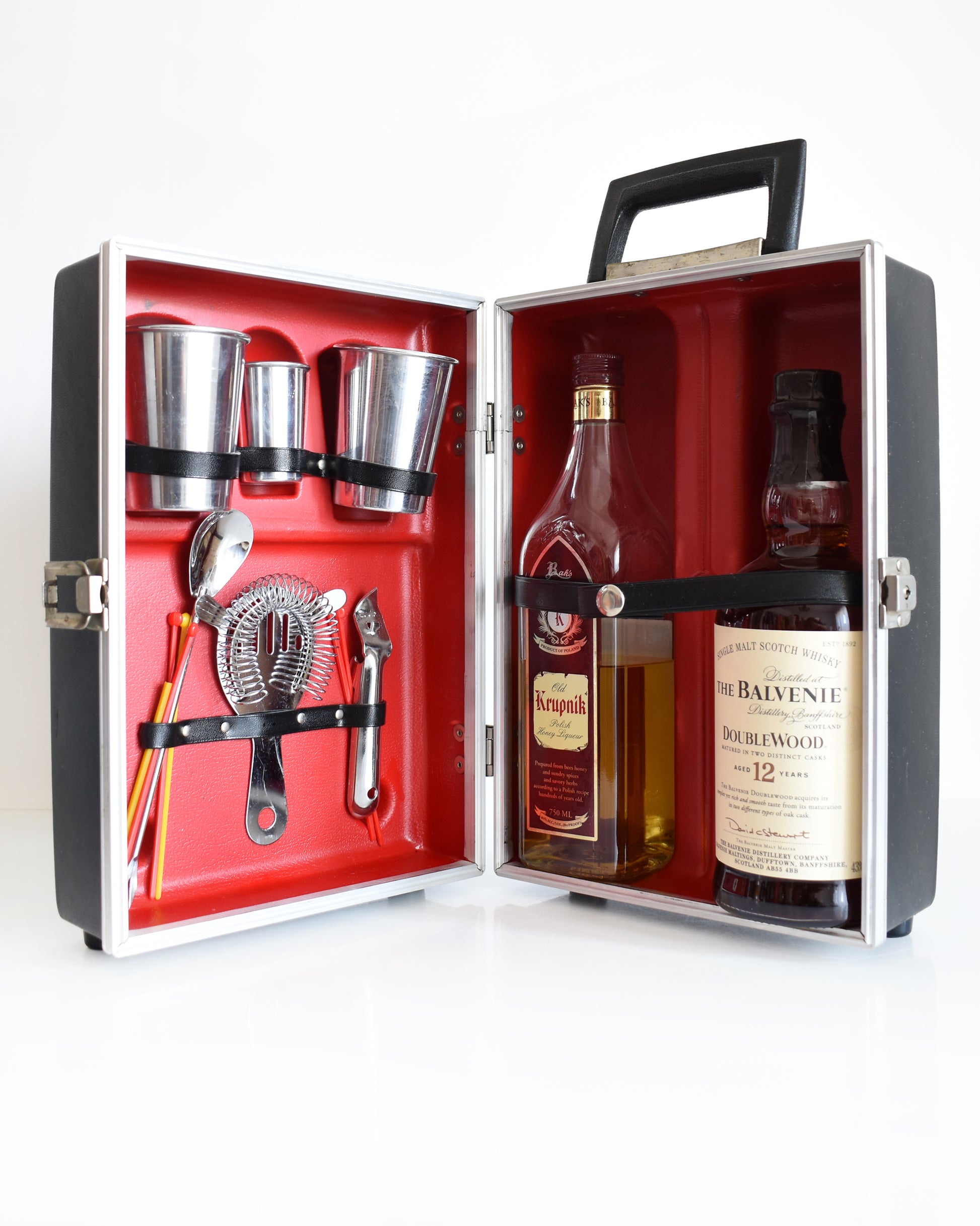 A vintage 1960s travel bar that is black and has a red molded inside to hold two aluminum glasses, one aluminum shot glass, a bottle opener, a drink strainer, a cocktail spoon/fork combo, and a few plastic stir sticks. There are two liquor bottles on the other side which aren't included.