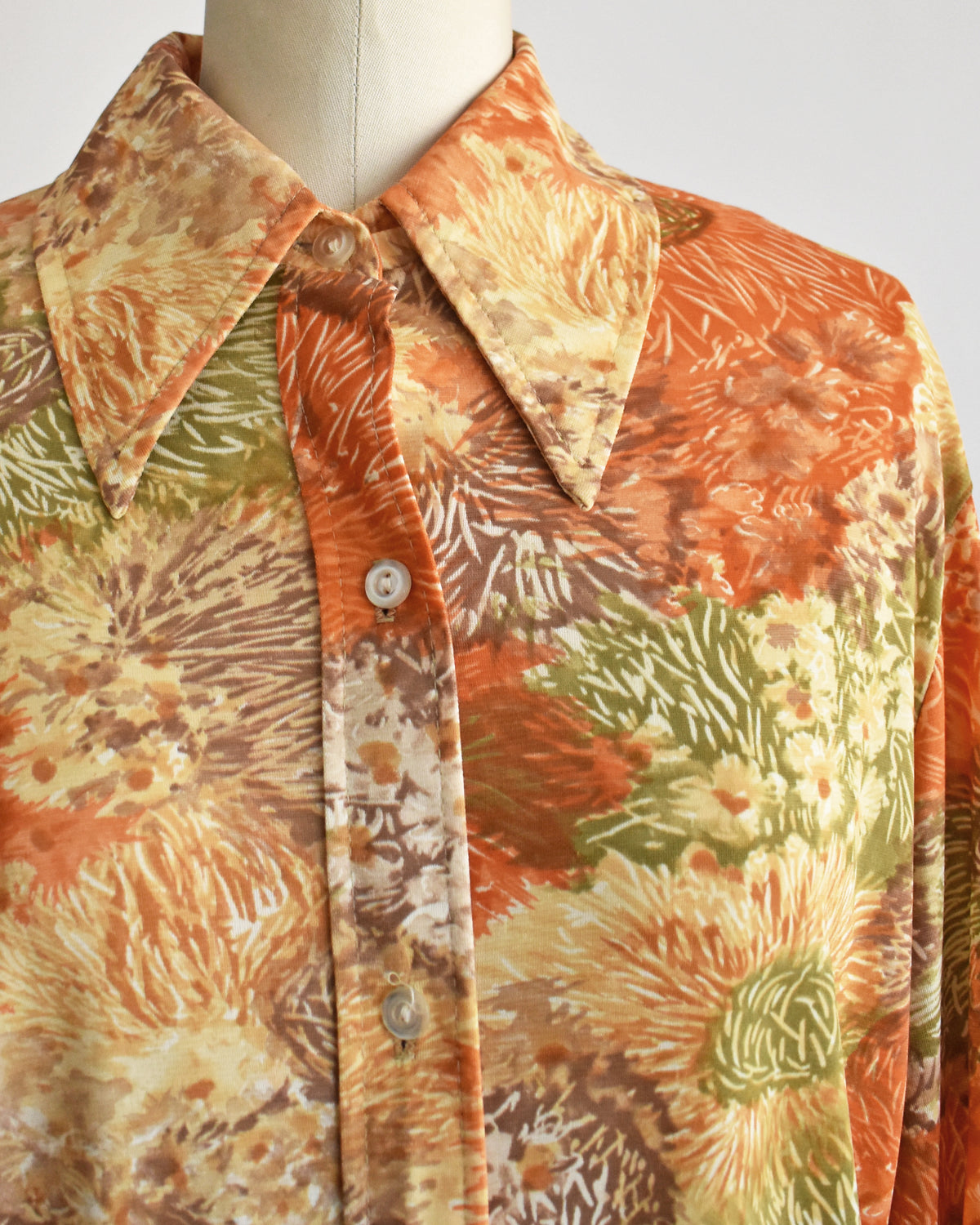 close up of a vintage 1970s long-sleeve button-down blouse which features an abstract floral watercolor print in orange, rust, and olive green tones displayed on a dress form.
