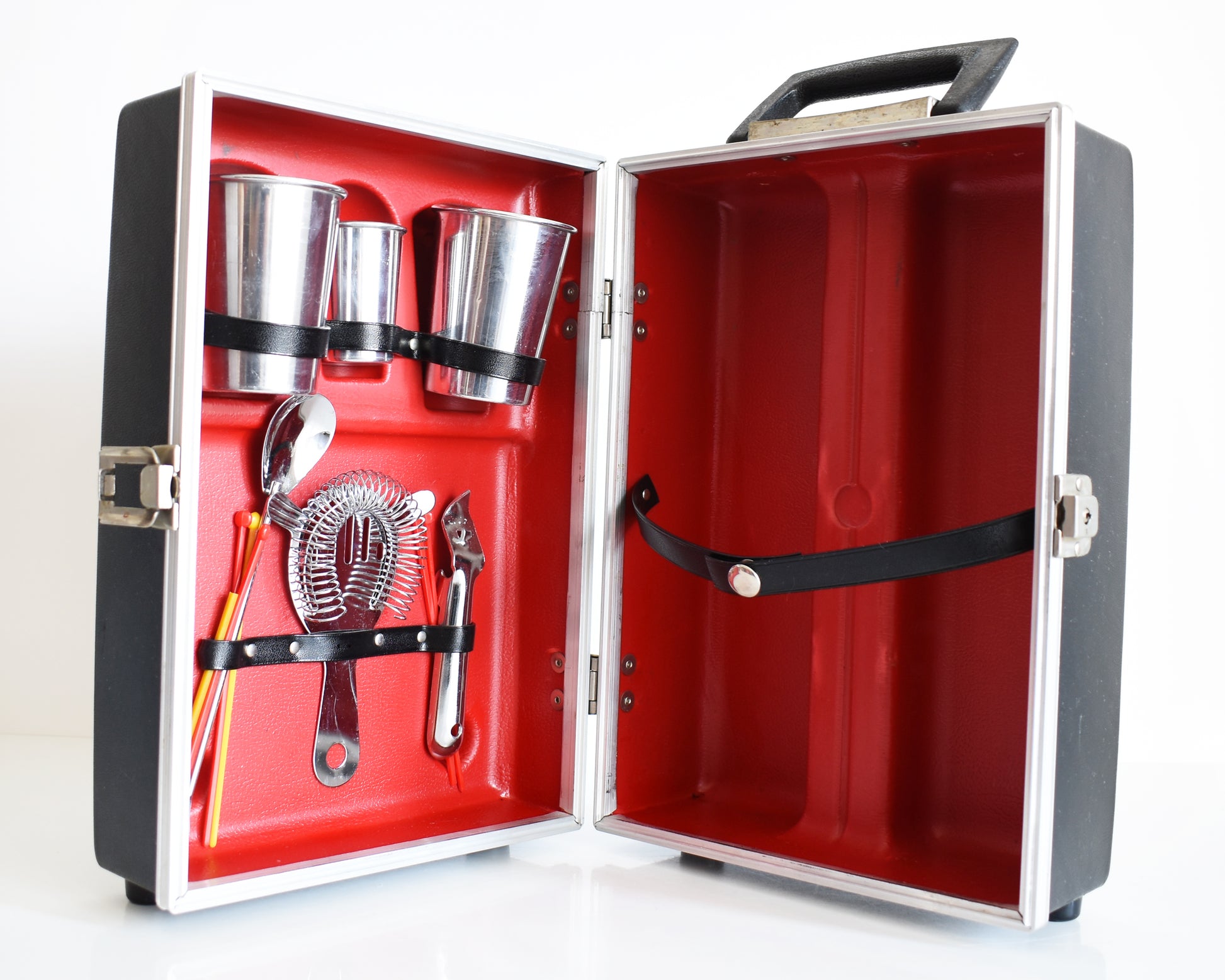 A vintage 1960s travel bar that is black and has a red molded inside to hold two aluminum glasses, one aluminum shot glass, a bottle opener, a drink strainer, a cocktail spoon/fork combo, and a few plastic stir sticks. The other side holds two bottles.