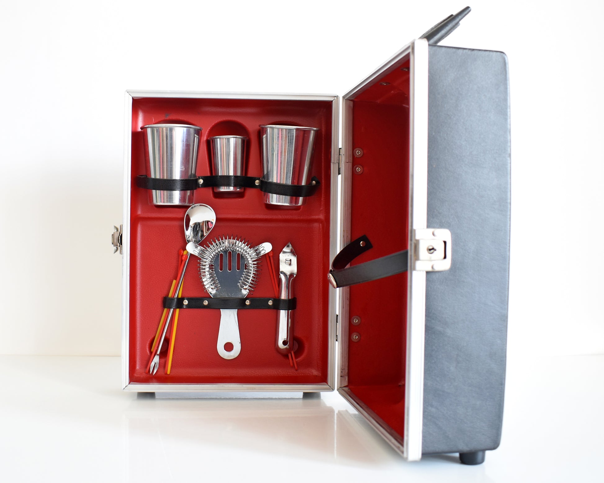 A vintage 1960s travel bar that is black and has a red molded inside to hold two aluminum glasses, one aluminum shot glass, a bottle opener, a drink strainer, a cocktail spoon/fork combo, and a few plastic stir sticks. 