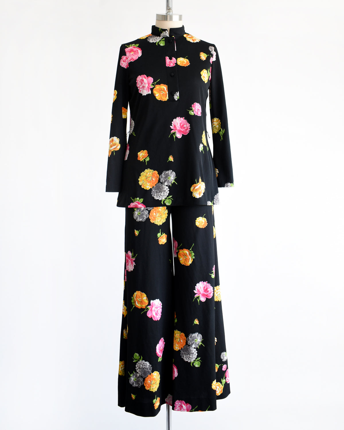 A vintage 1970s black two-piece suit set with a colorful floral print, displayed on a mannequin against a plain white background. The set includes a long-sleeve mock-neck top and matching wide-leg trousers.