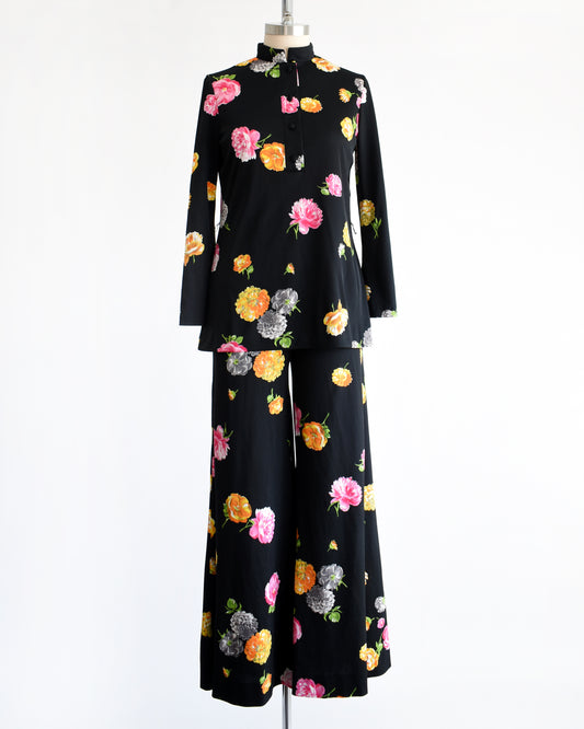 A vintage 1970s black two-piece suit set with a colorful floral print, displayed on a mannequin against a plain white background. The set includes a long-sleeve mock-neck top and matching wide-leg trousers.