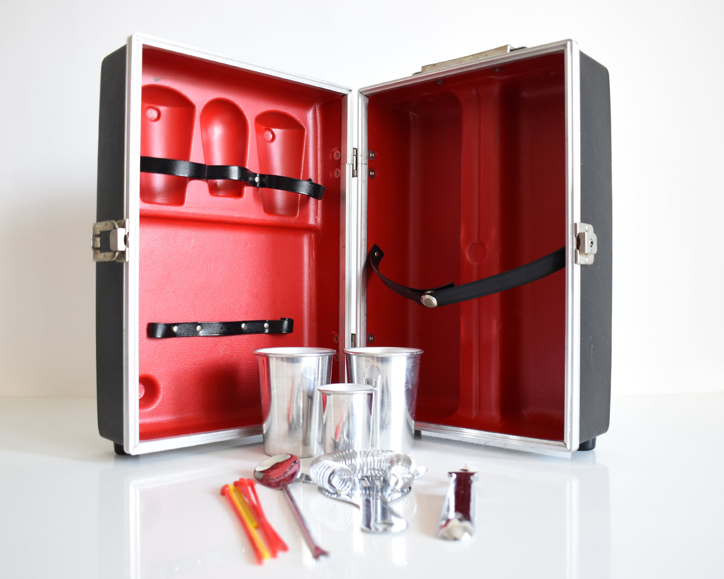 A vintage 1960s travel bar that is black and has a red molded inside to hold two aluminum glasses, one aluminum shot glass, a bottle opener, a drink strainer, a cocktail spoon/fork combo, and a few plastic stir sticks. The glasses and utensils  are outside the case showing the molded slots.