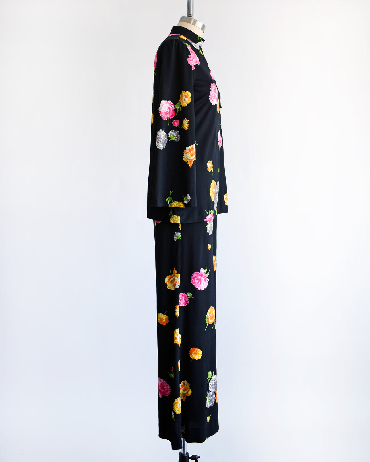 side view of a vintage 1970s black two-piece suit set with a colorful floral print, displayed on a mannequin against a plain white background. The set includes a long-sleeve mock-neck top and matching wide-leg trousers.
