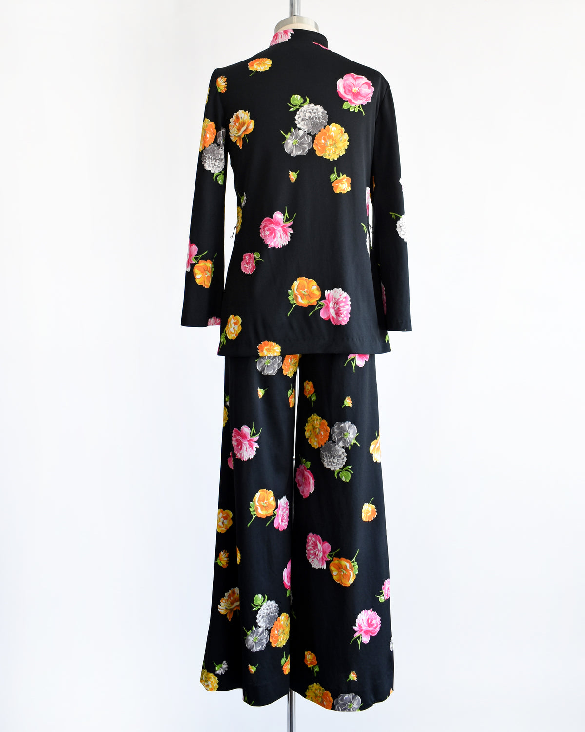 back view of a vintage 1970s black two-piece suit set with a colorful floral print, displayed on a mannequin against a plain white background. The set includes a long-sleeve mock-neck top and matching wide-leg trousers.