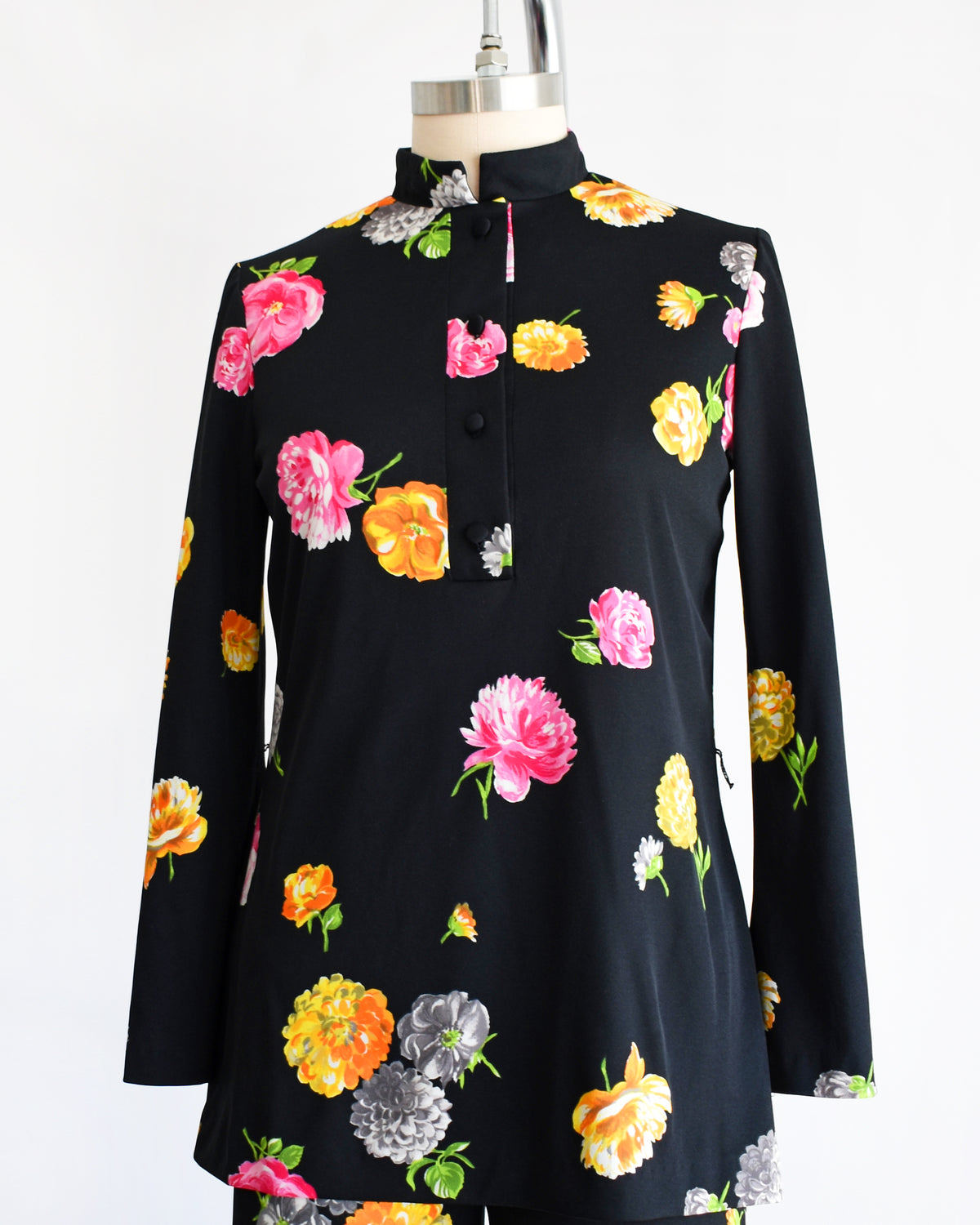 side front. view of a vintage 1970s black two-piece suit set with a colorful floral print, displayed on a mannequin against a plain white background. The set includes a long-sleeve mock-neck top and matching wide-leg trousers.