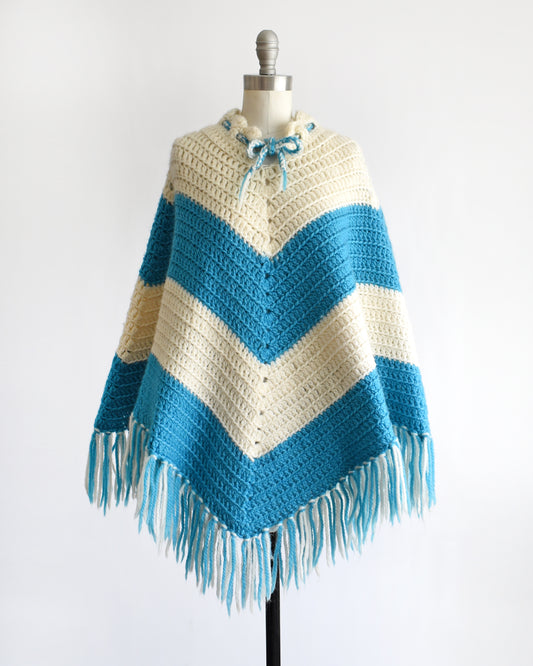 a vintage 1970s blue and white chevron striped fringed poncho on a dress form against a white background.