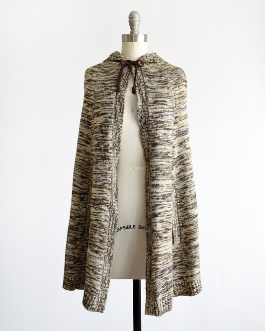 a vintage 1970s hooded poncho with a marled knit texture in beige, brown, and black, displayed on a cream dress form mannequin against a white background.