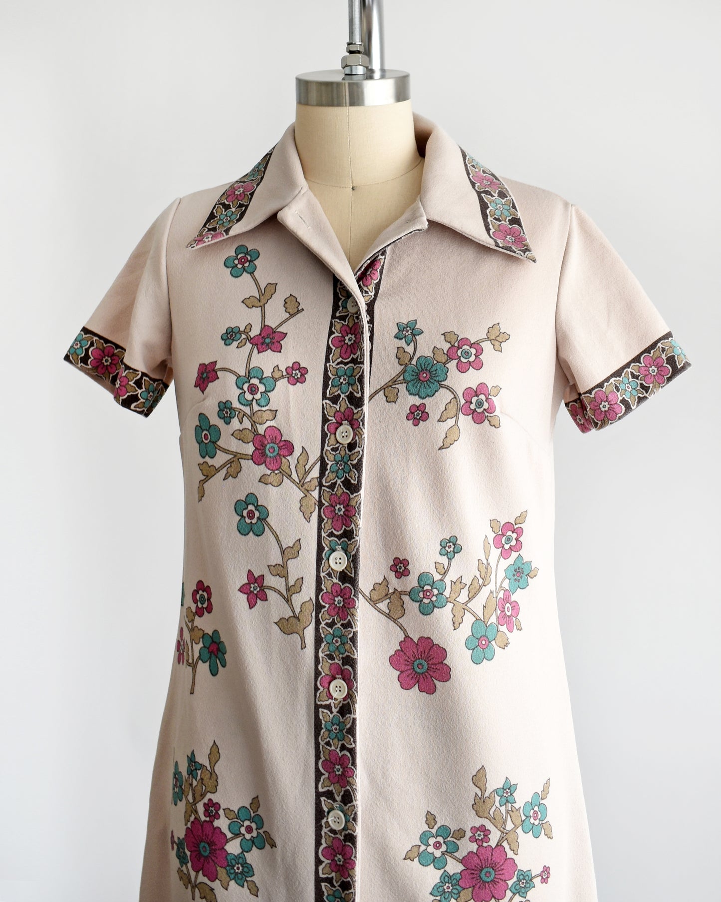 A vintage 1970s light brown mod dress with button front that features a blue and pink floral print. The top button is unbuttoned in this photo