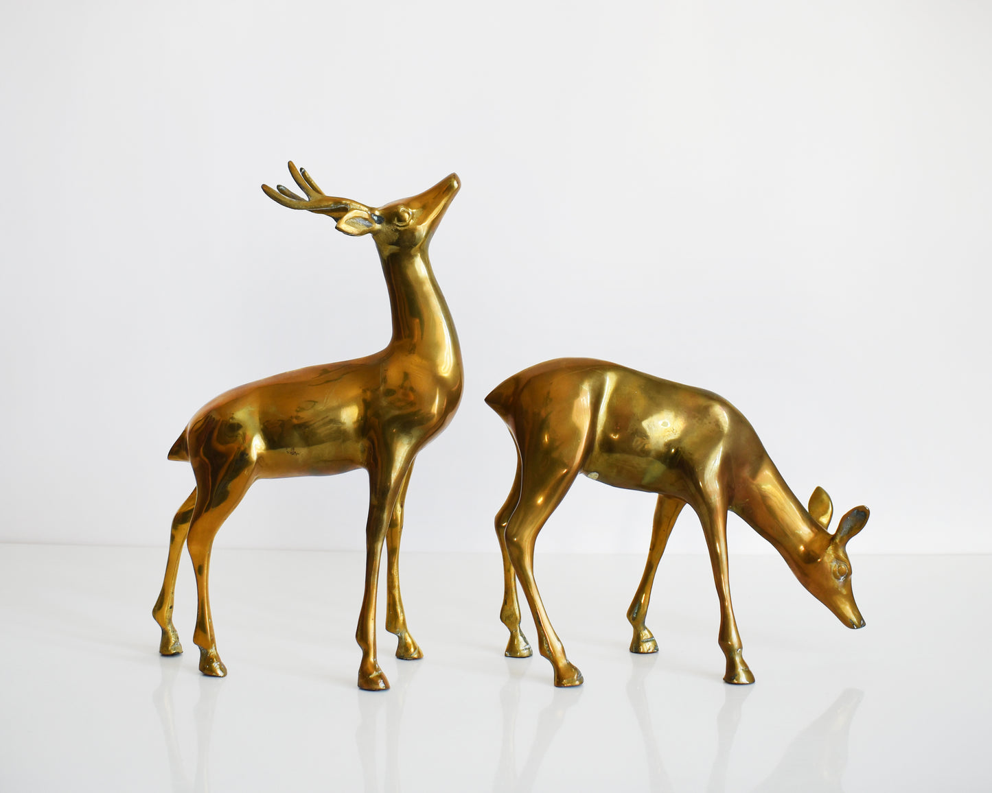 A pair of two large vintage brass deer figurines. One buck and one doe make up this collection, with the buck standing upright displaying his antlers and the doe is grazing on the ground.