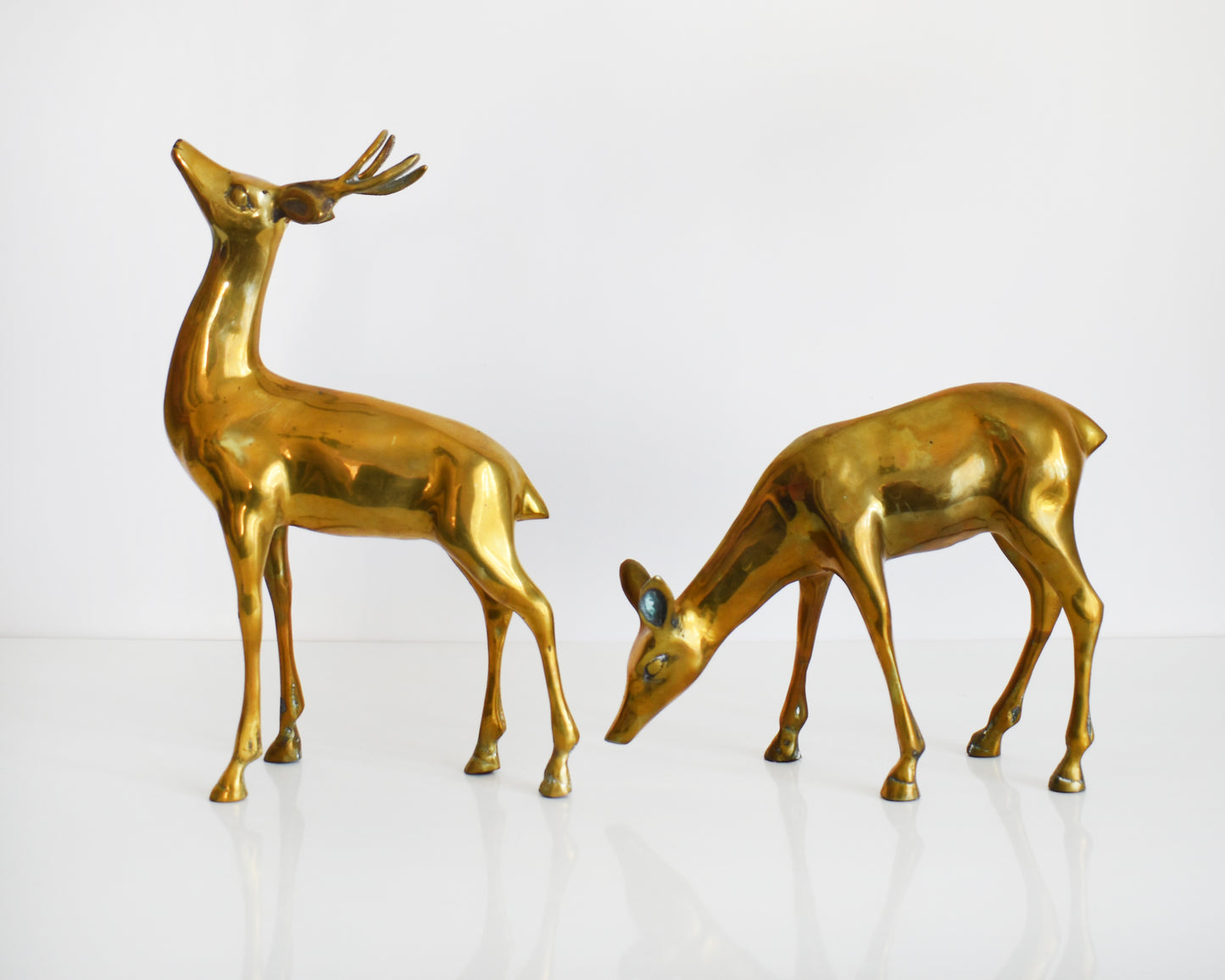 A pair of two large vintage brass deer figurines. One buck and one doe make up this collection, with the buck standing upright displaying his antlers and the doe is grazing on the ground.
