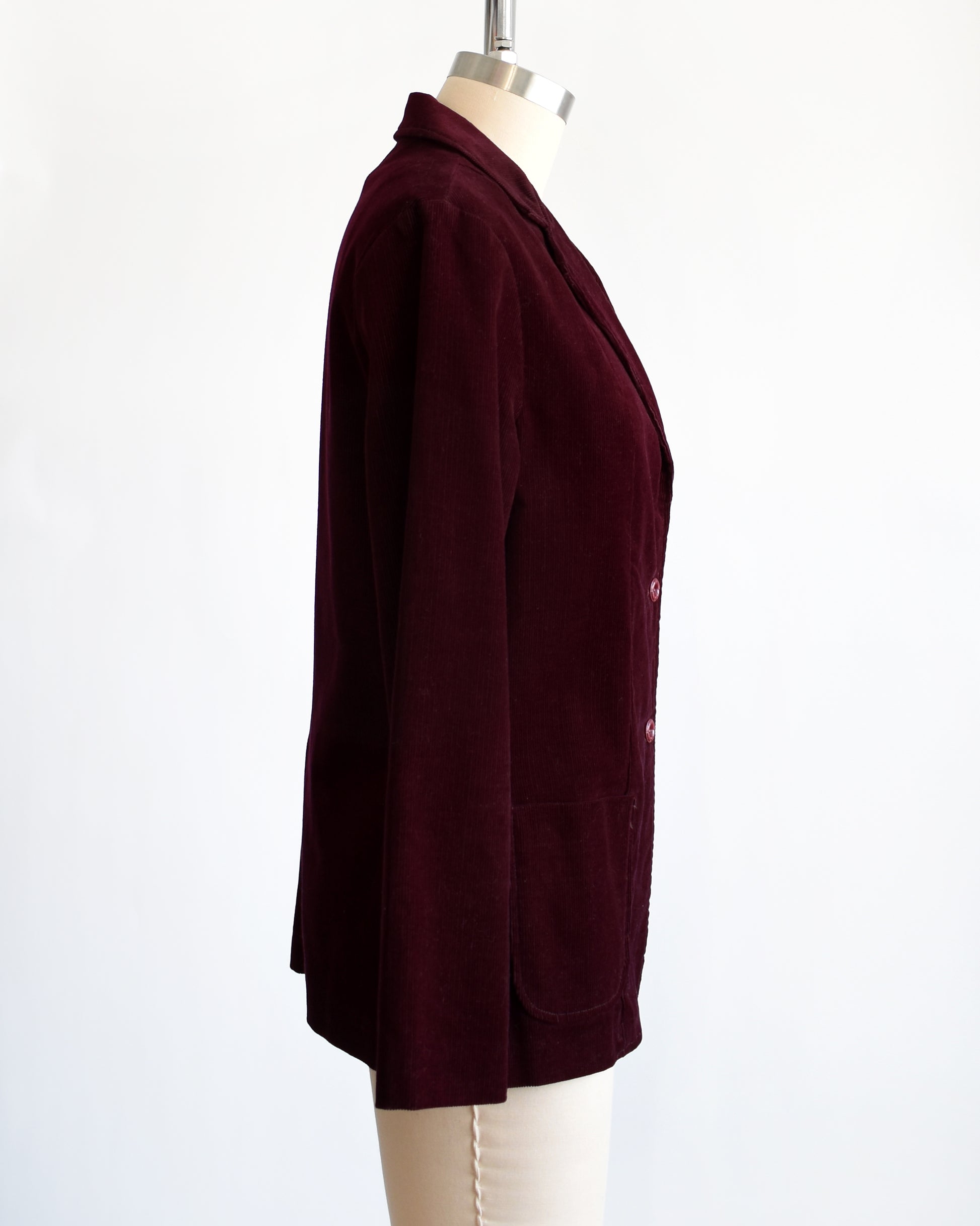 Side view of a vintage 1980s burgundy corduroy blazer features two plastic buttons on the front and two front patch pockets on each side.