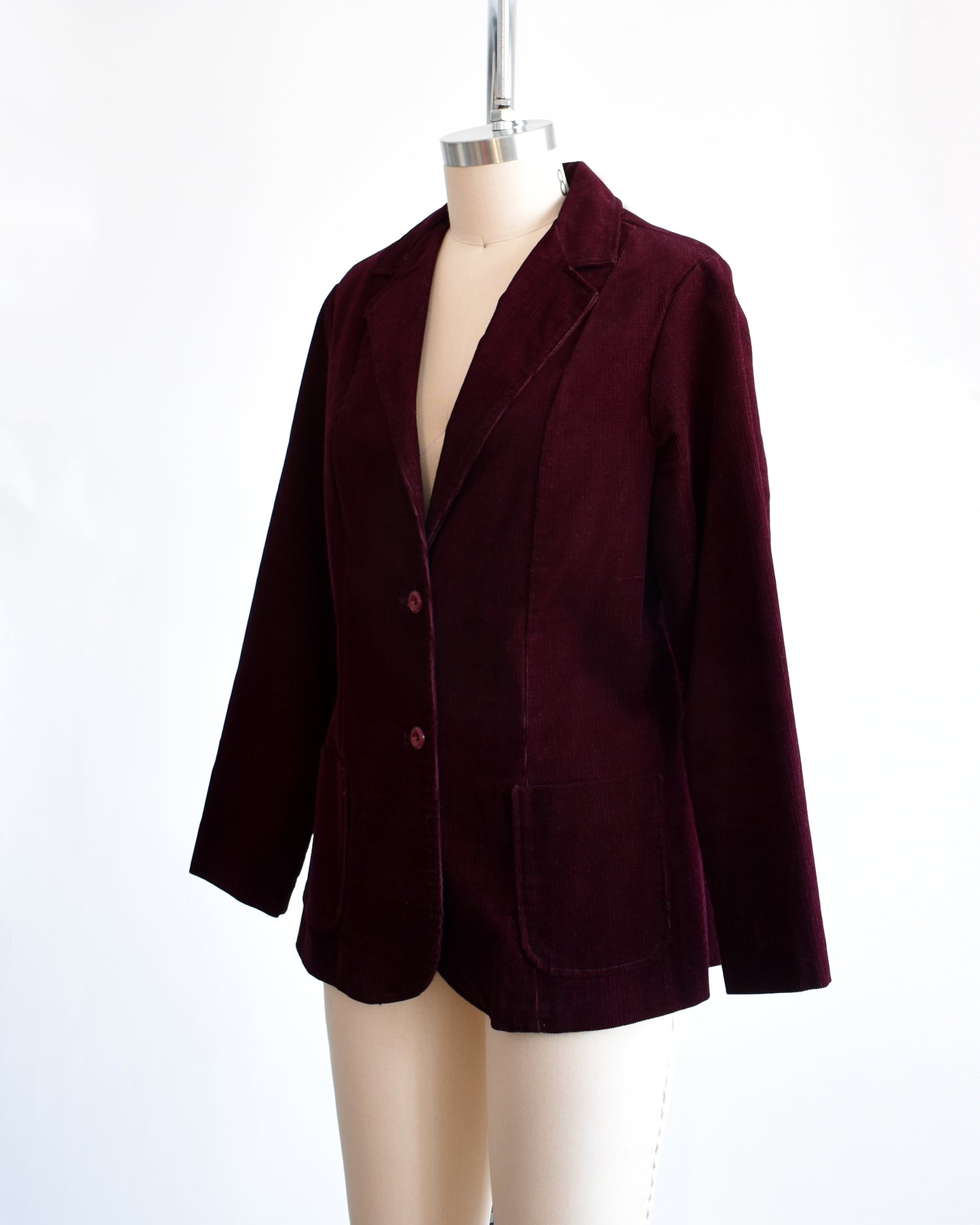 Side front view of  a vintage 1980s burgundy corduroy blazer features two plastic buttons on the front and two front patch pockets on each side.