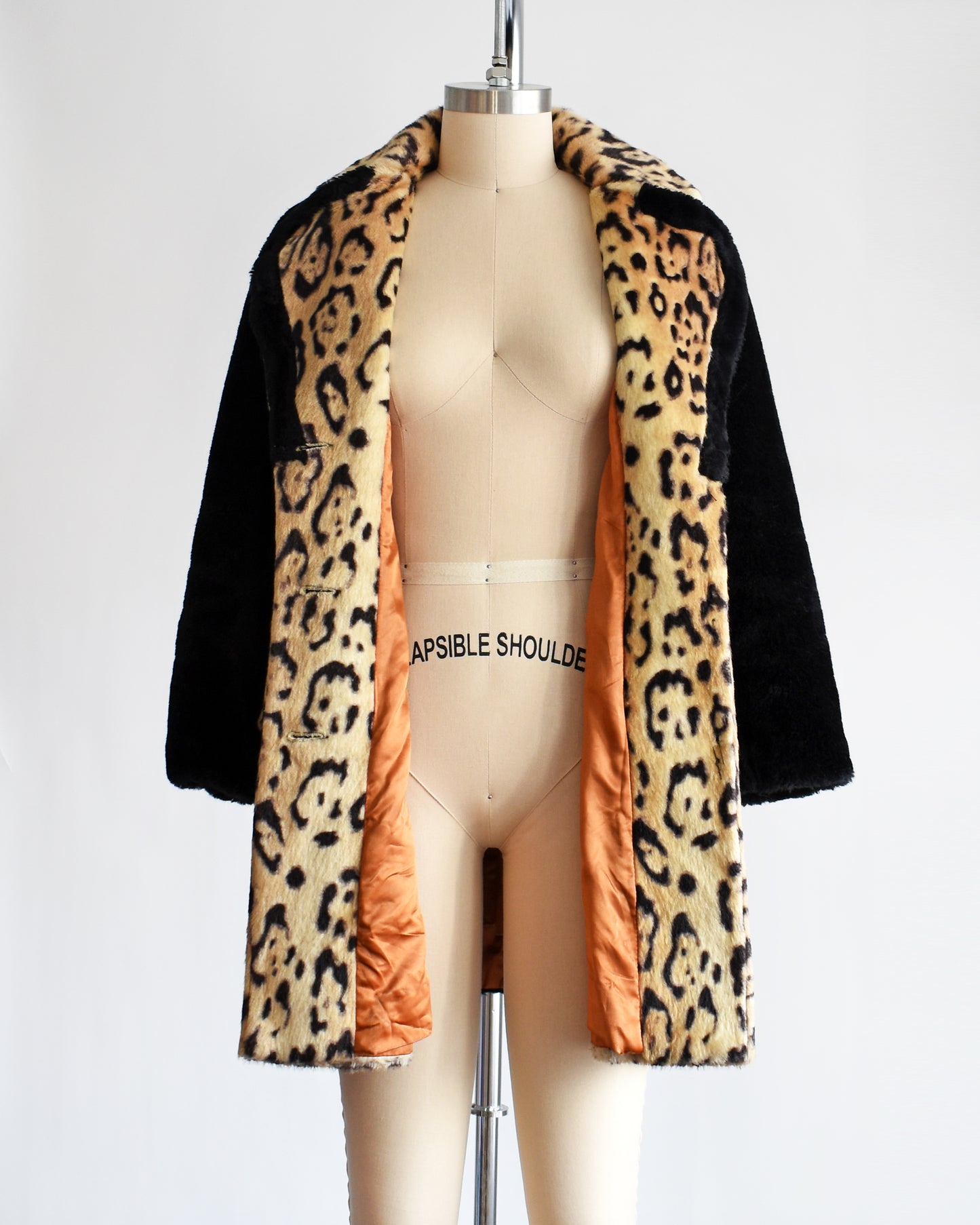 A vintage late 1960s early 1970s leopard faux fur coat that has large leopard print on the collar, down the front, and on the large side pockets. Black plush long sleeves, sides, and back. Matching black trim on the collar. The coat is opened showing the orange lining.