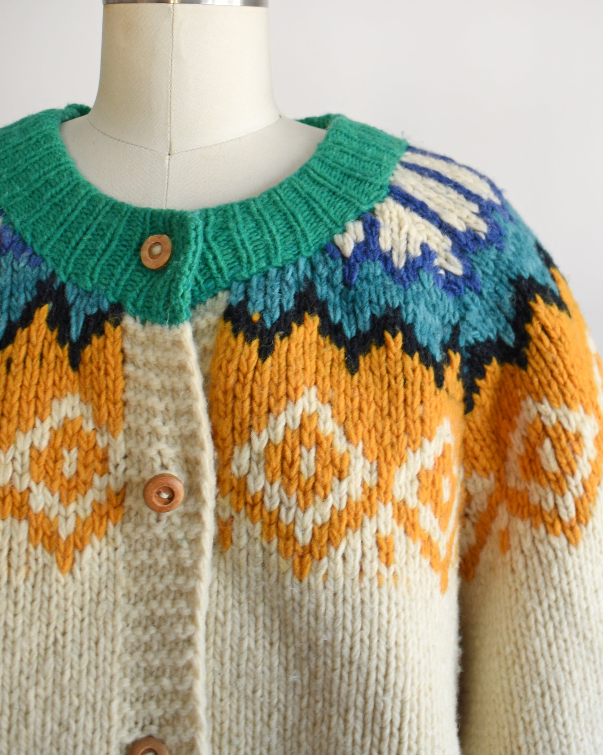 Close up of a vintage 1970s cream wool cardigan with bright green, dark blue, light blue, black, cream, and light orange Fair Isle pattern around the collar, and wood buttons on the front.