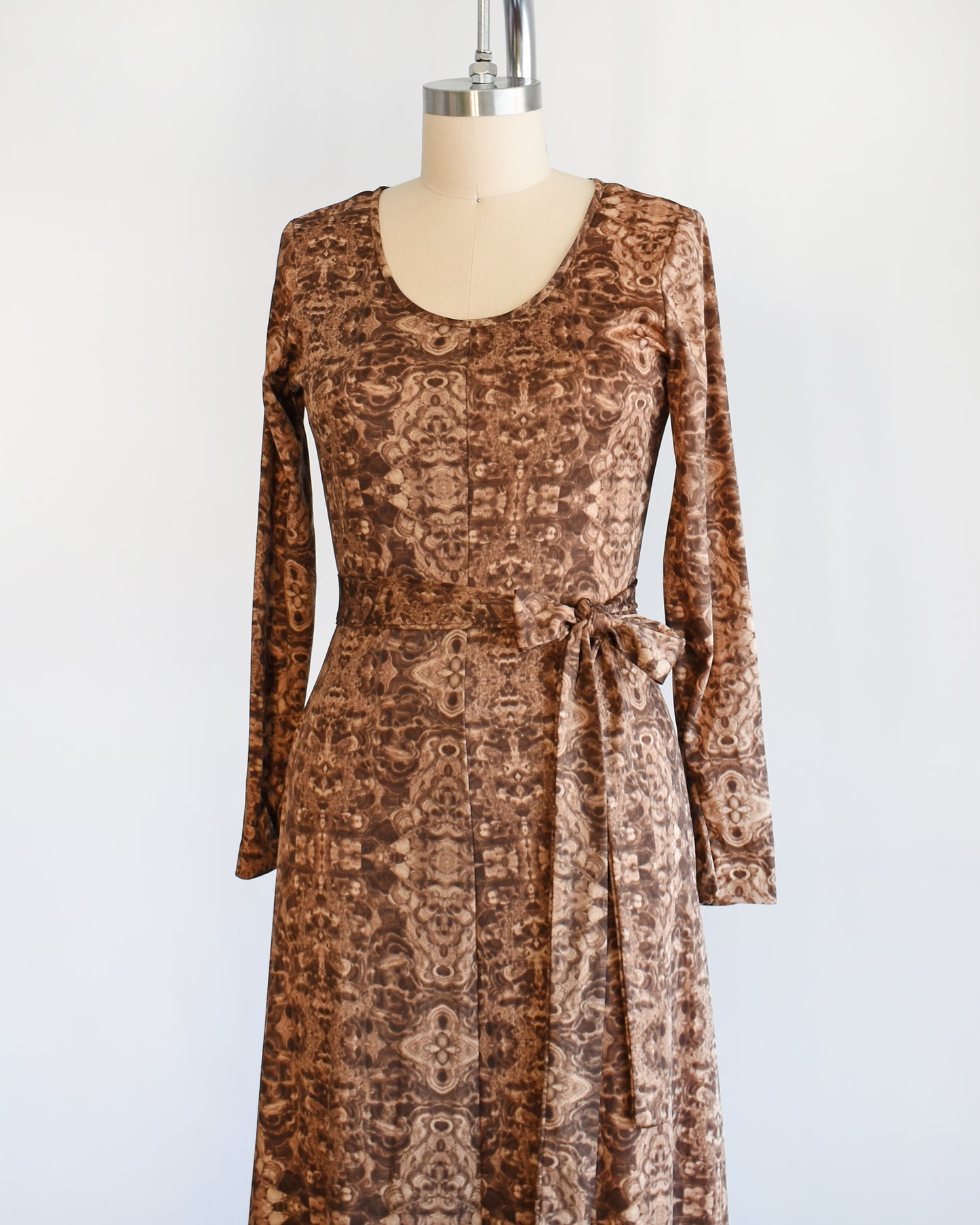 Side front view of a vintage 1970s long sleeve maxi dress which features a psychedelic kaleidoscope pattern in light and dark brown.