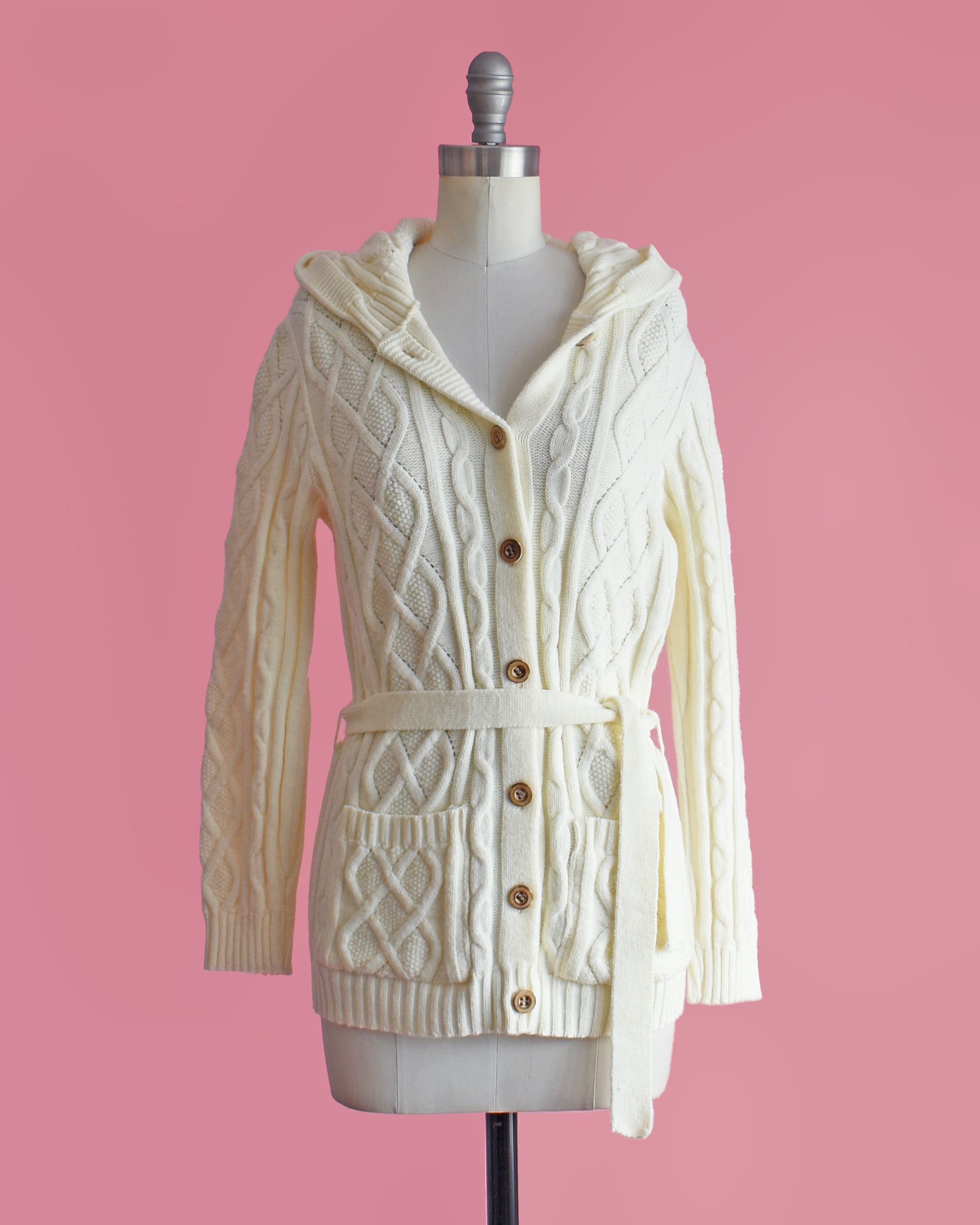 a vintage 1970s cream-colored cable knit hooded cardigan on a mannequin against a pink background