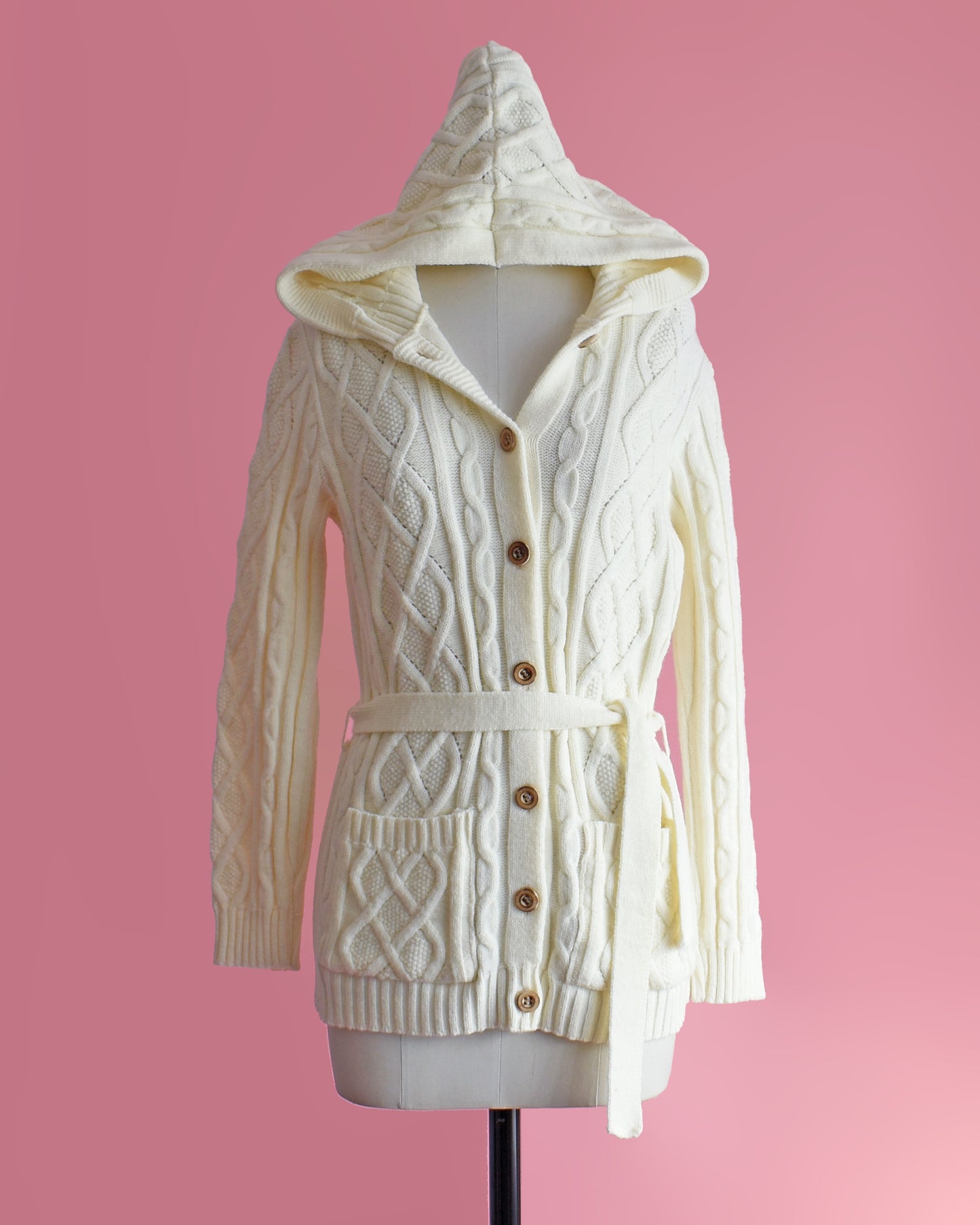 a vintage 1970s cream-colored cable knit hooded cardigan on a mannequin against a pink background. The hood is pulled forward in this photo