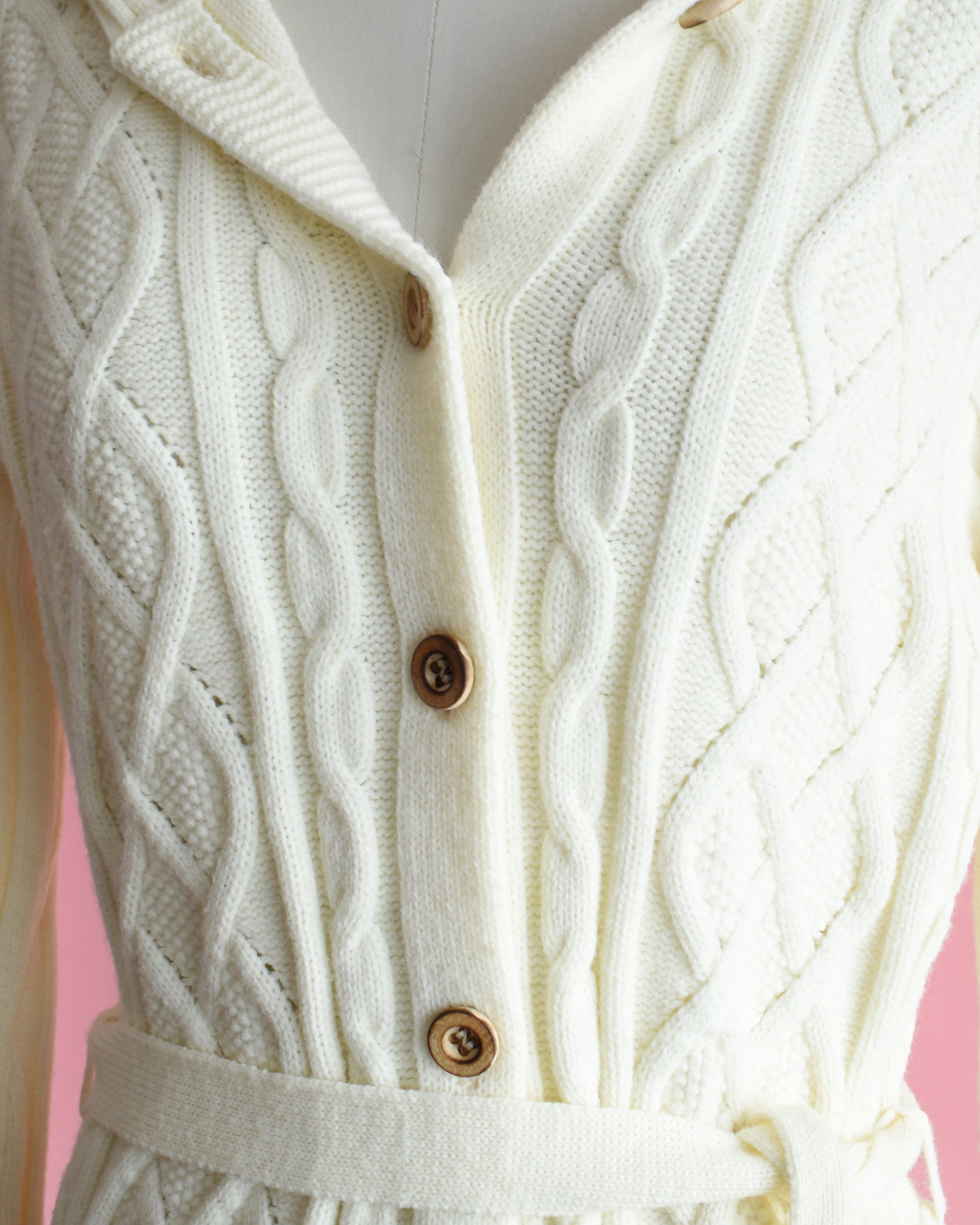 close up of the wood buttons on a vintage 1970s cream-colored cable knit hooded cardigan on a mannequin against a pink background