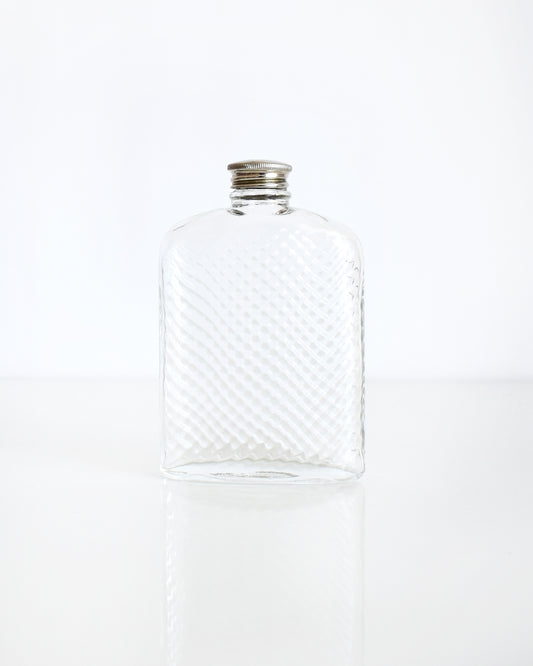 A vintage glass flask that has a cut diamond pattern and a silver screw top. The flask is on a white table and a white background.